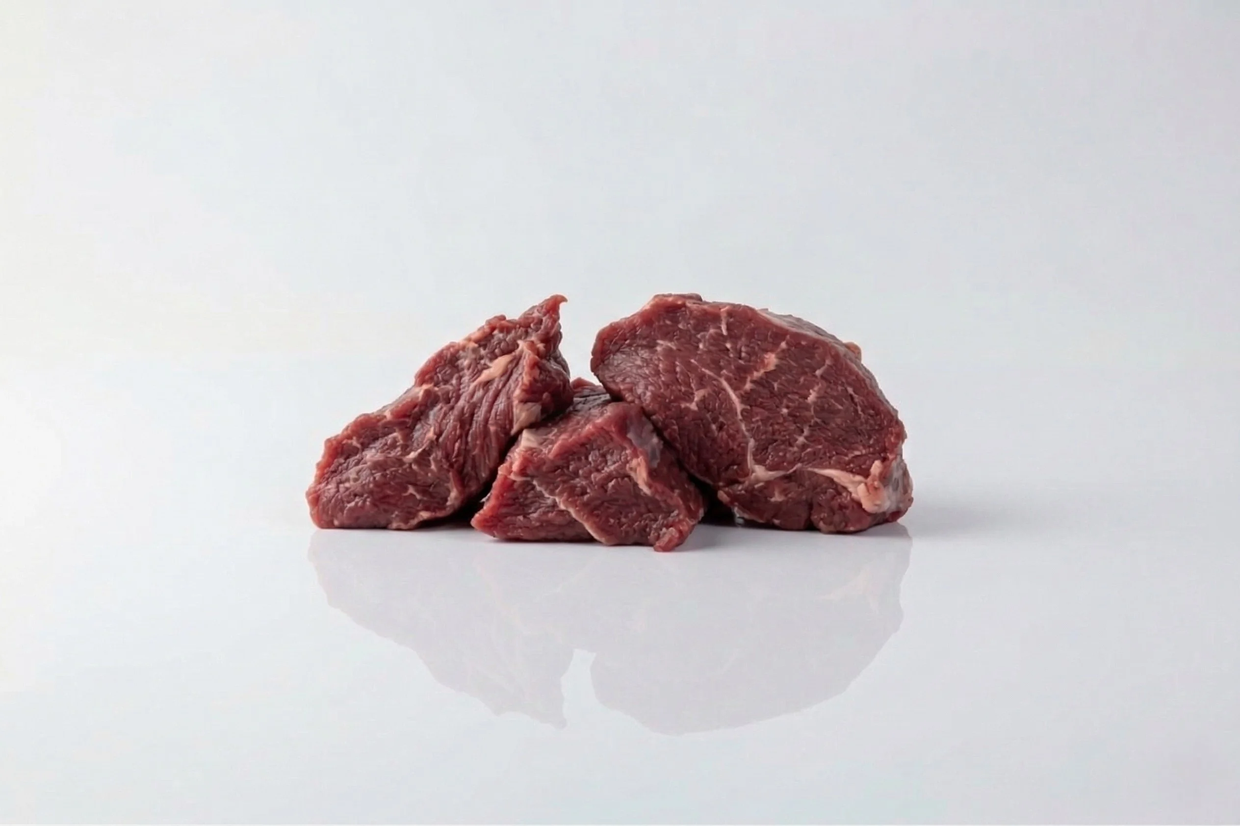 A photograph of three raw pieces of goat stew meat with visible marbling placed on a white reflective surface against a light grey background.