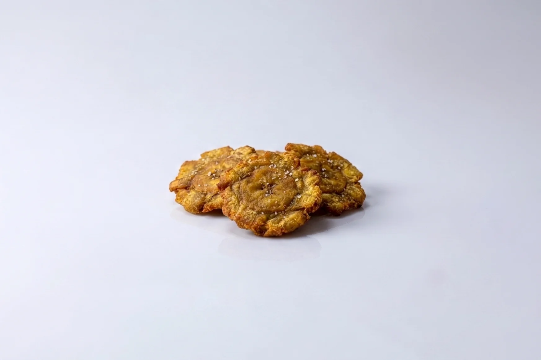 Three golden-fried tostones (twice-fried flattened plantains) sprinkled with salt on a white reflective surface against a light grey background.