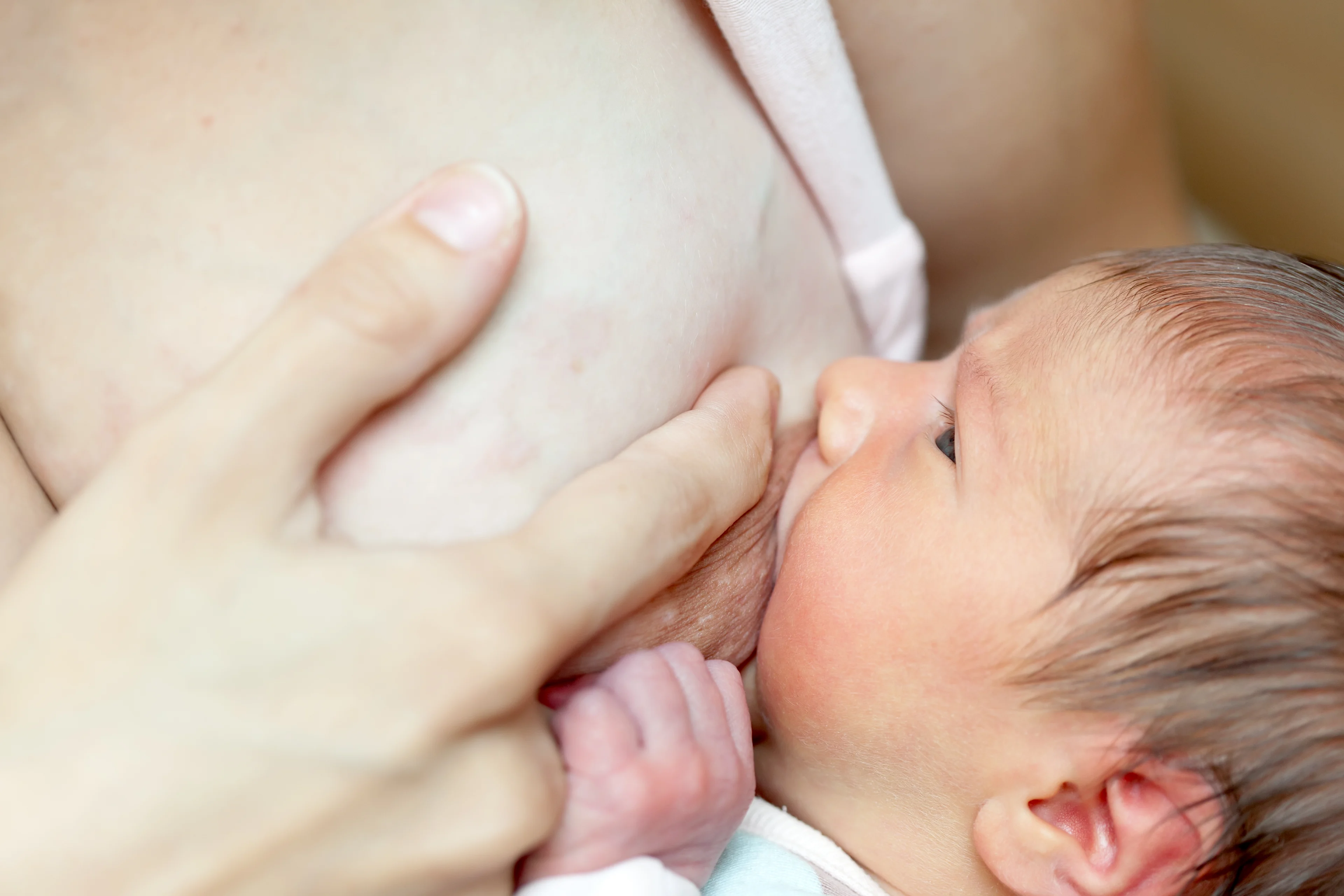 Alt text: A close-up of a newborn breastfeeding, with the mother's hand supporting her breast and the baby's tiny fist visible.