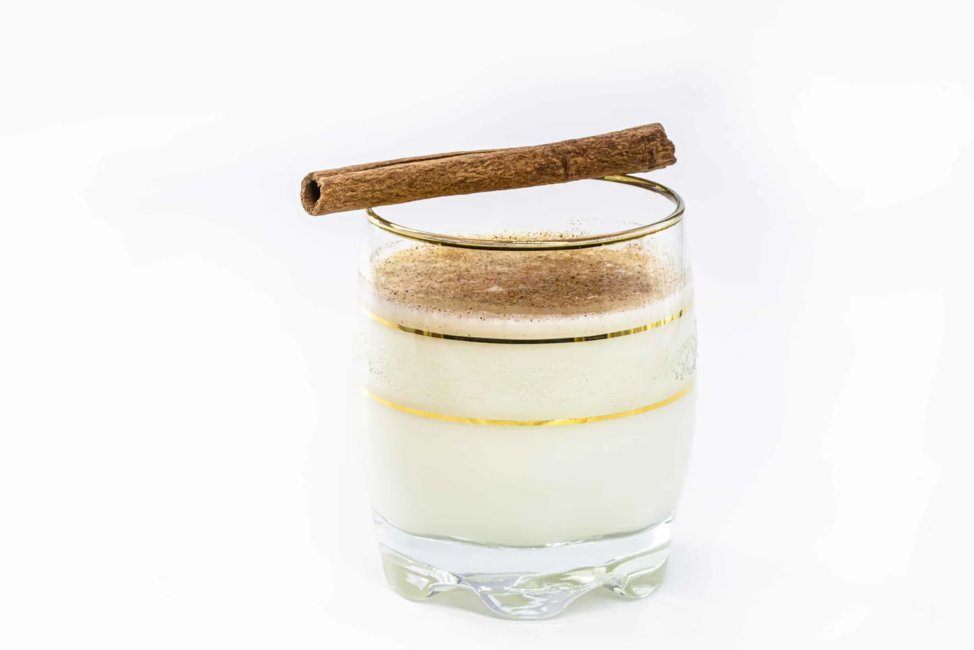 a small round glass with thin gold stripes filled with a white liquid, dusted with a brown spice, with a cinnamon stick laid on top of the glass