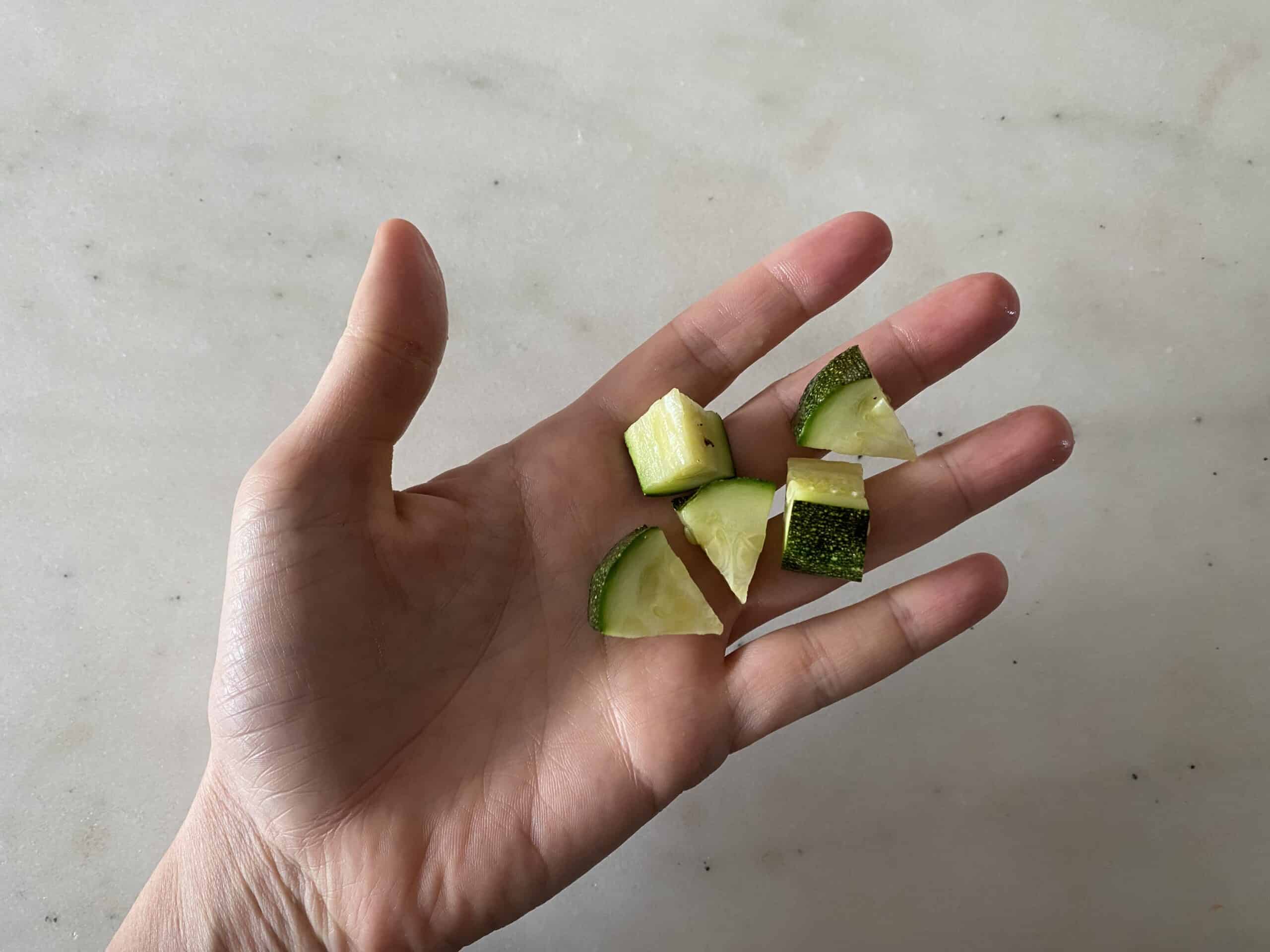 a hand holding five bite-sized pieces of cooked zucchini for babies 9 months+