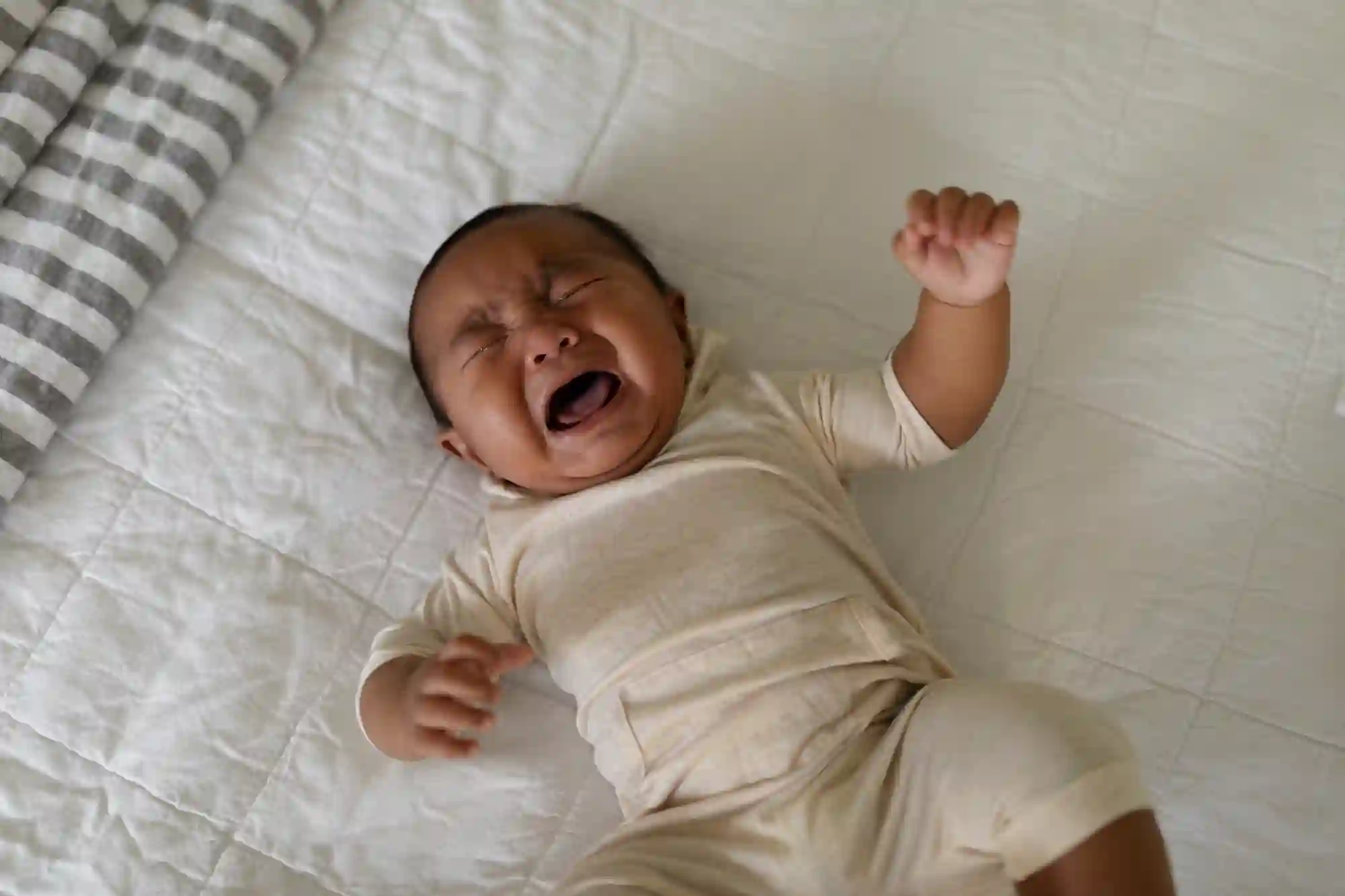 A baby in a cream-colored onesie lying on a white quilted surface, crying with eyes shut and fists raised.