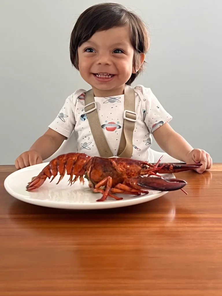 smiling toddler sitting at a wooden table in front of a plate containing a whole lobster