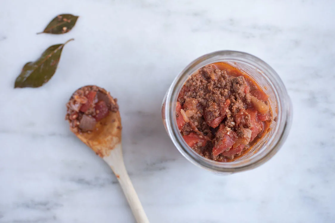 a glass jar filled with bolognese sauce next to a wooden spoon and two bay leaves on a marble background