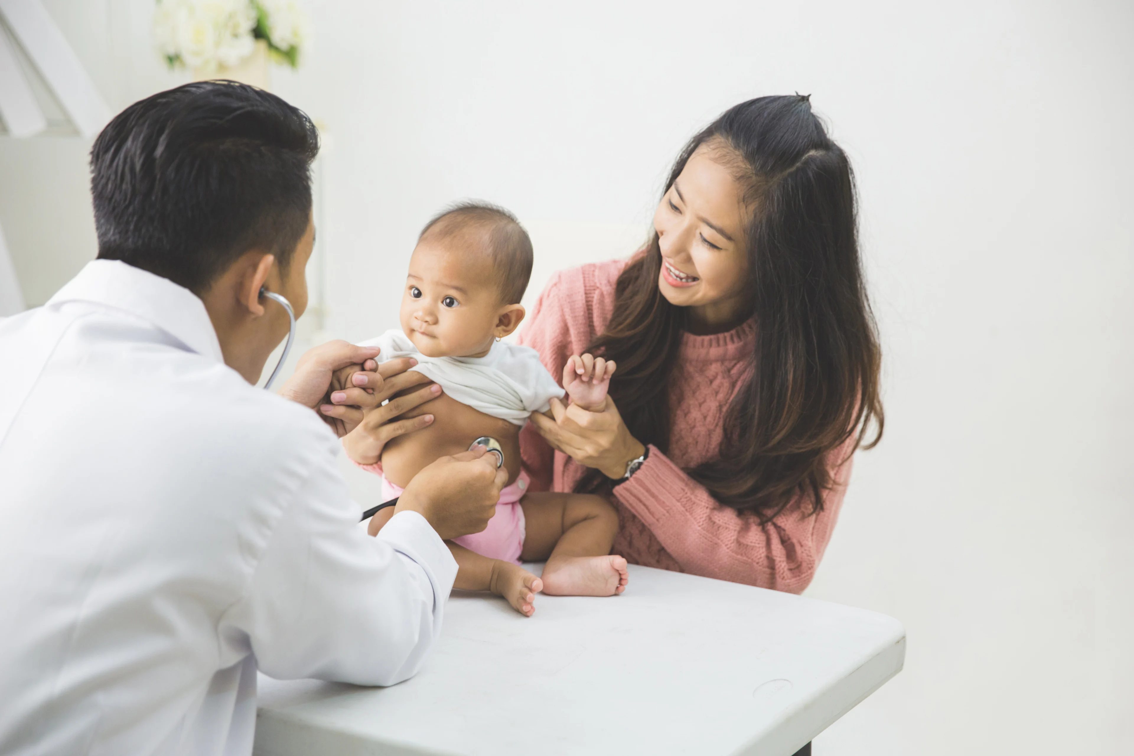 A doctor with a stethoscope examines a baby held by a smiling woman in a pink sweater. The setting is bright and clinical, conveying a caring atmosphere.