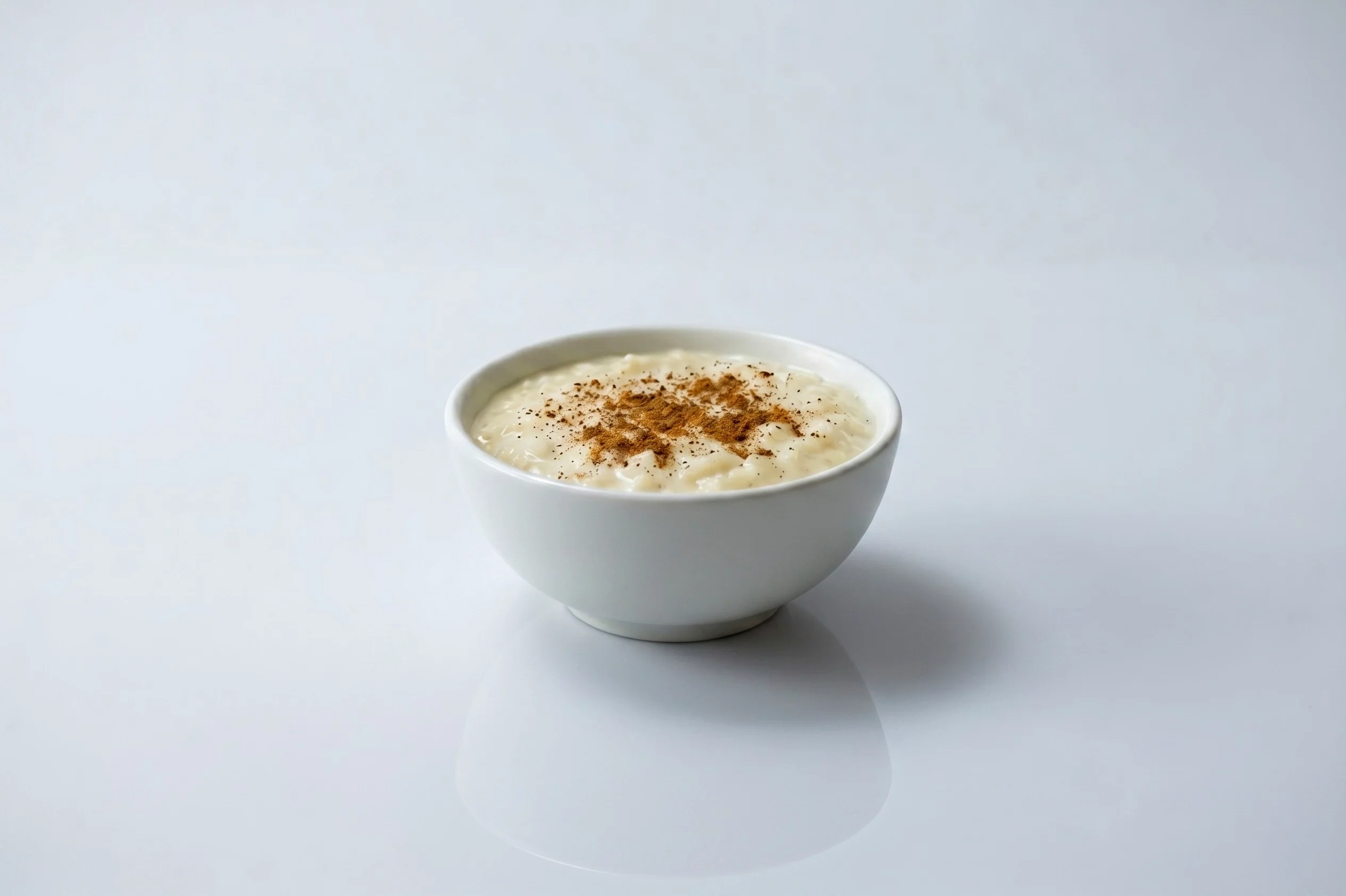 A white ceramic bowl of creamy arroz con leche (rice pudding) topped with a sprinkle of cinnamon, on a white reflective surface against a light grey background.