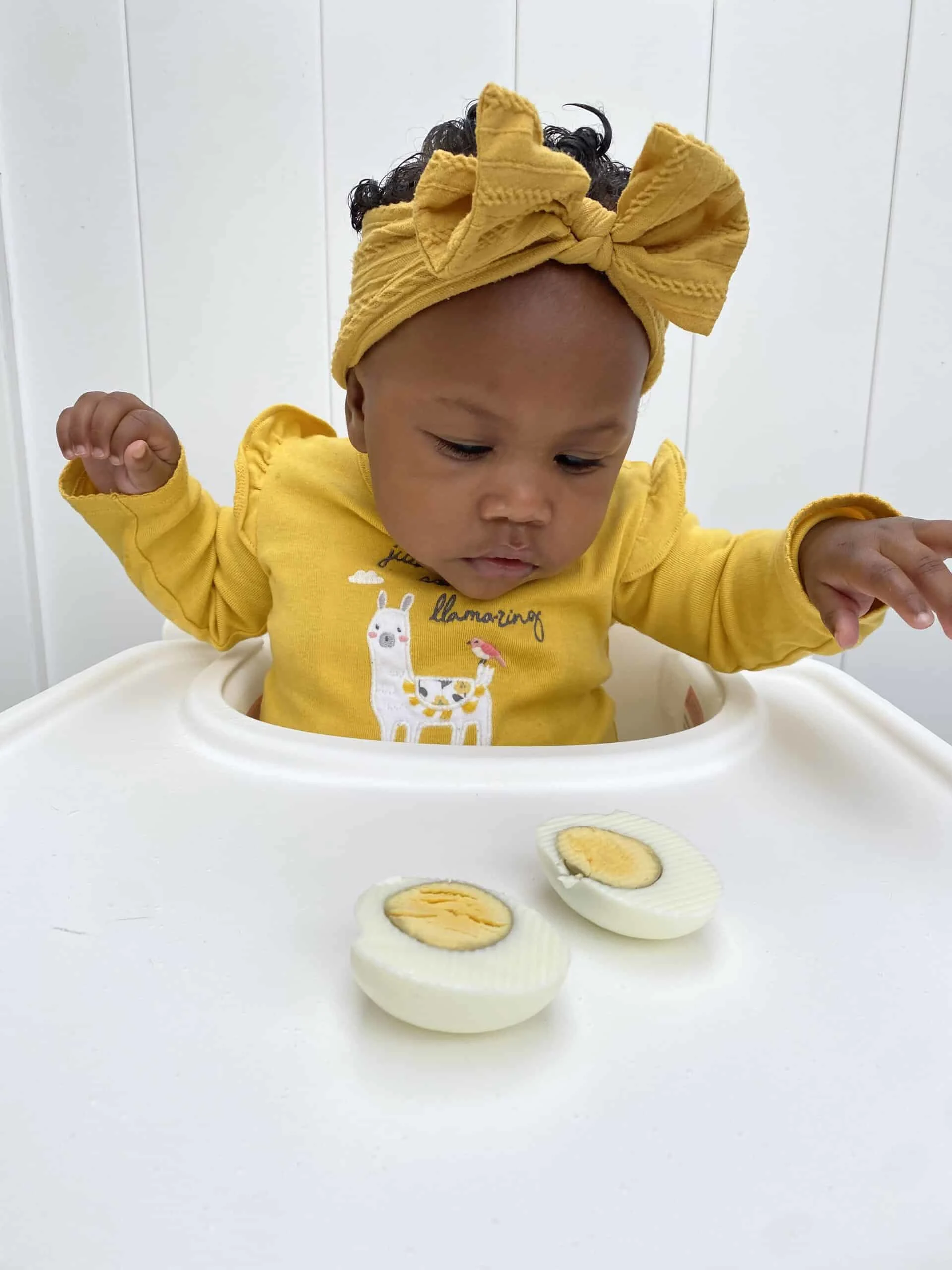 a baby wearing a yellow shirt and headband looks at a hard boiled egg cut in half
