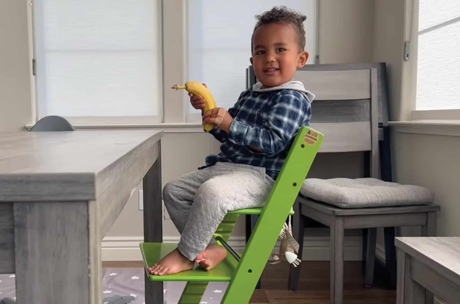 toddler smiling and sitting on a green high chair holding a banana with his hands