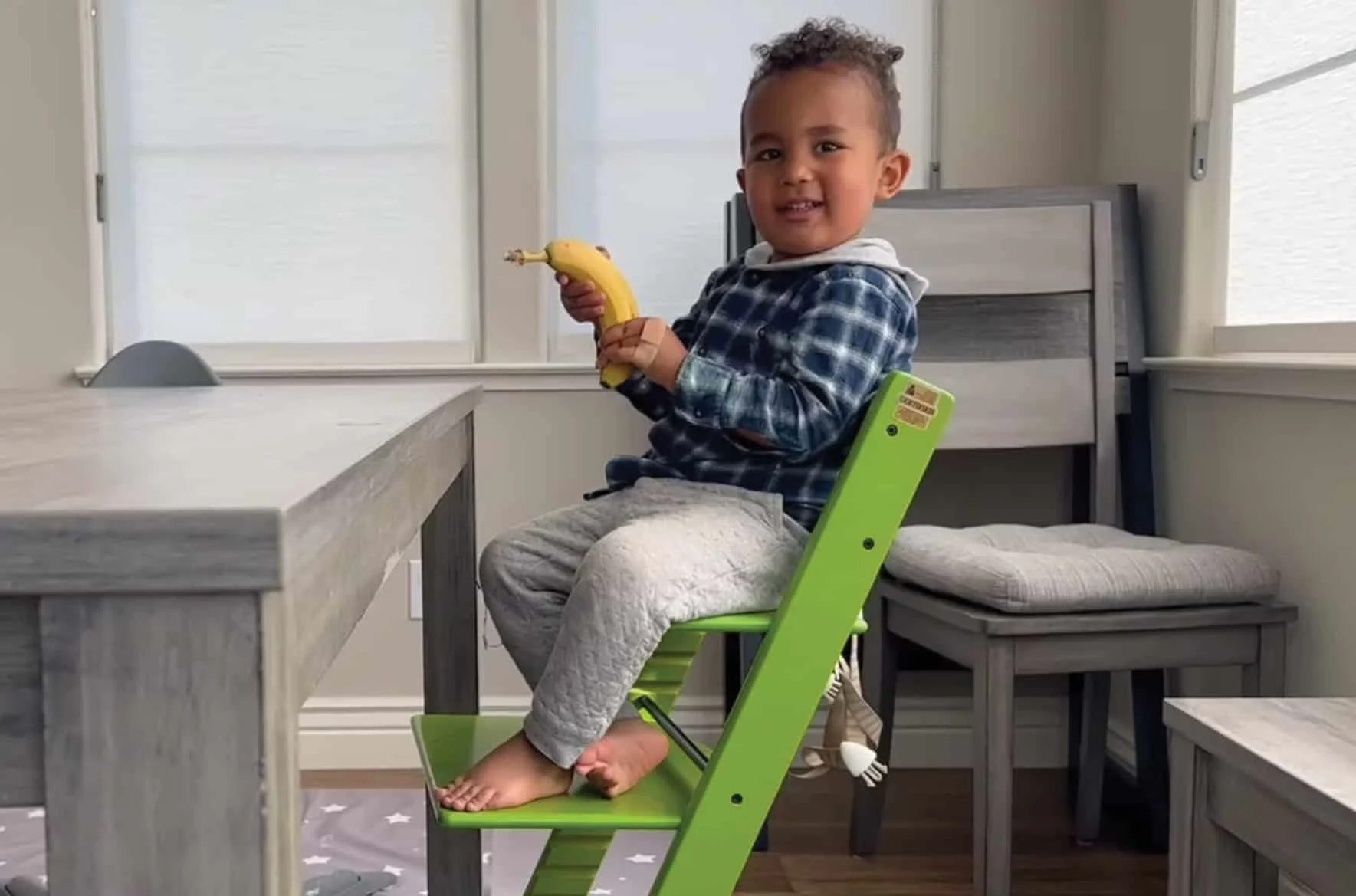 un niño pequeño soriendo sentado en una silla alta verde y sosteniendo una banana en sus manos
