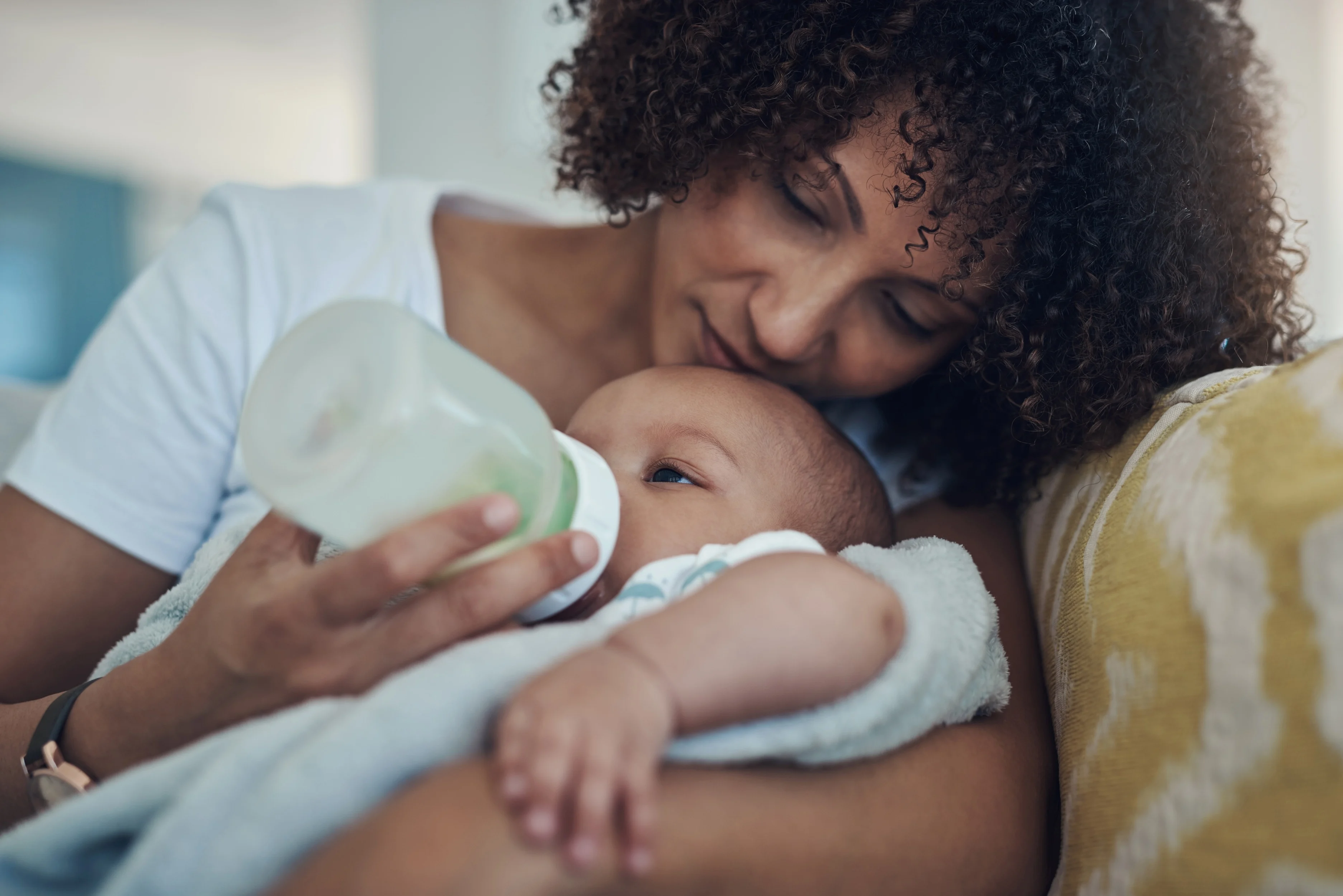 A mother with curly hair tenderly resting her cheek on her baby's head while bottle-feeding the infant on a couch.