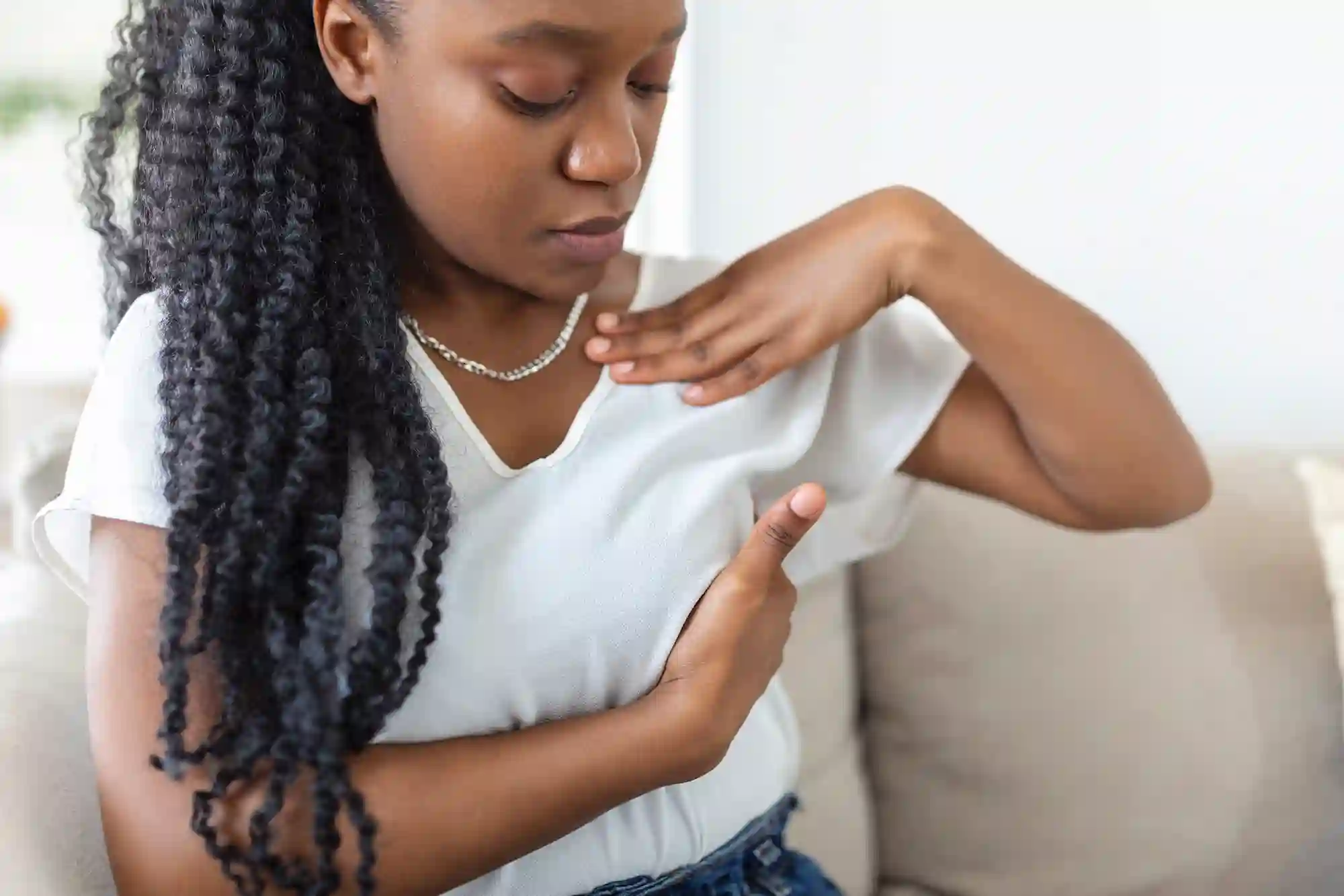 A young Black woman with long curly hair sits on a couch, looking down and touching her chest area, suggesting breast pain or discomfort.