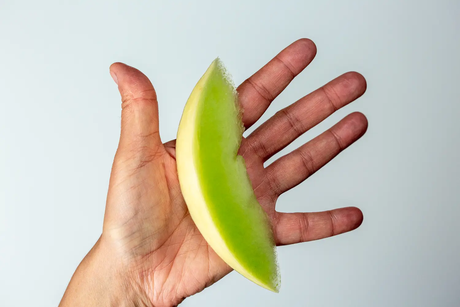 a hand holding a wedge of honeydew melon with the rind on 