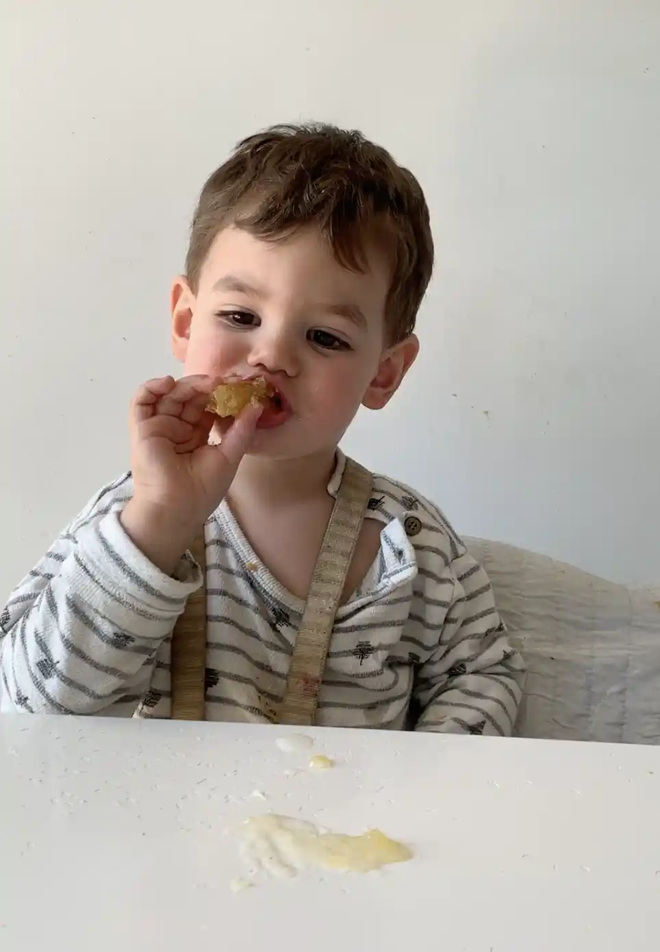 Toddler holding a piece of honey to his mouth, while sitting in a high chair in front of a white table with splatters of honey on it