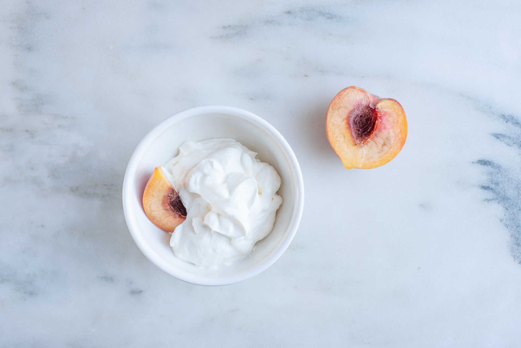a white bowl filled with whipped cream and a soft ripe peach half next to another peach half on a marble background