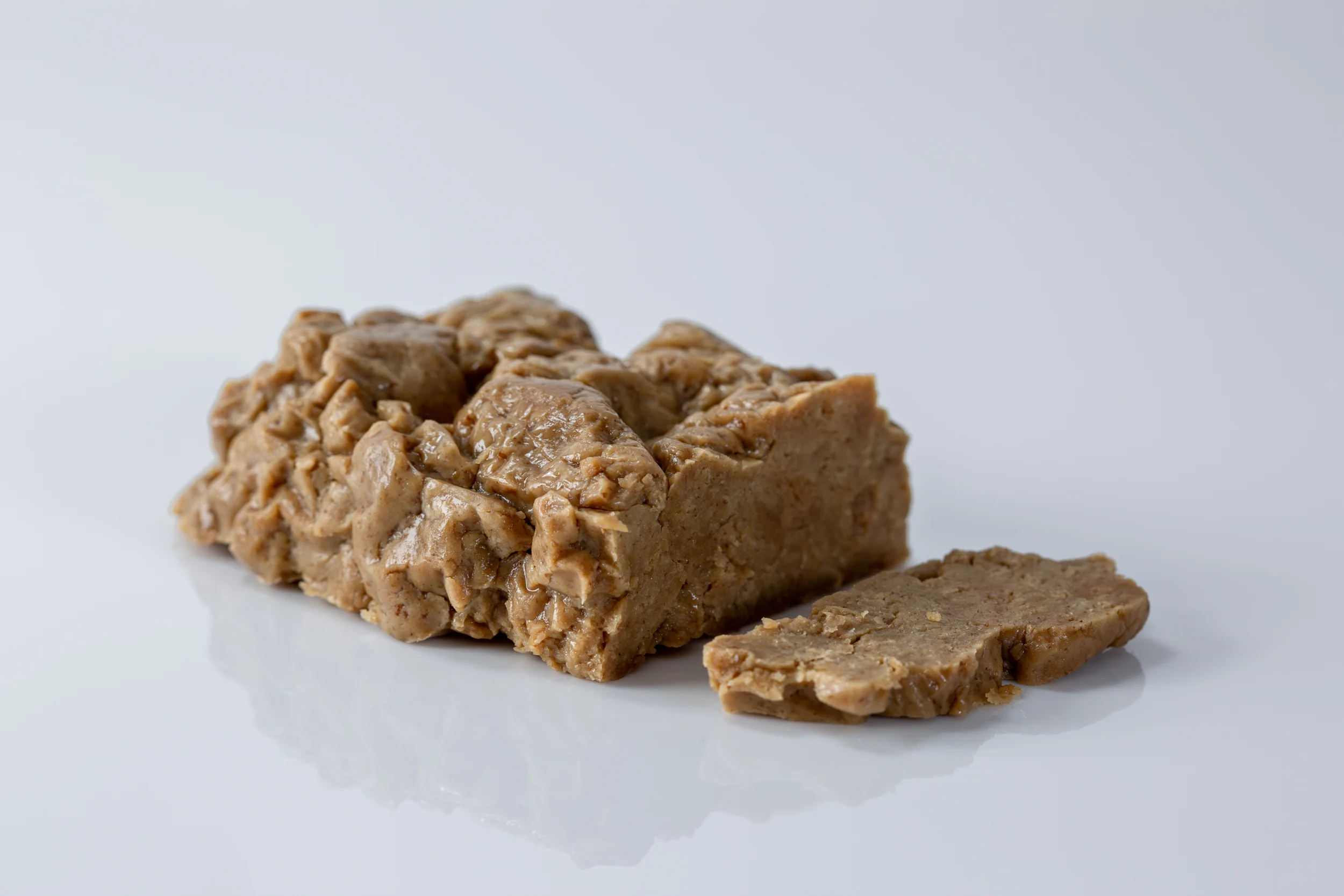 A block of seitan with a rough, textured surface on a white reflective surface, with one slice cut and placed beside it, revealing a dense, firm interior.