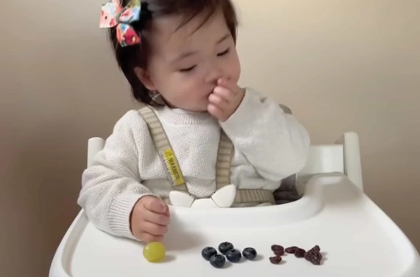 toddler sitting on a white high chair with a tray, eating blueberries and a green grape