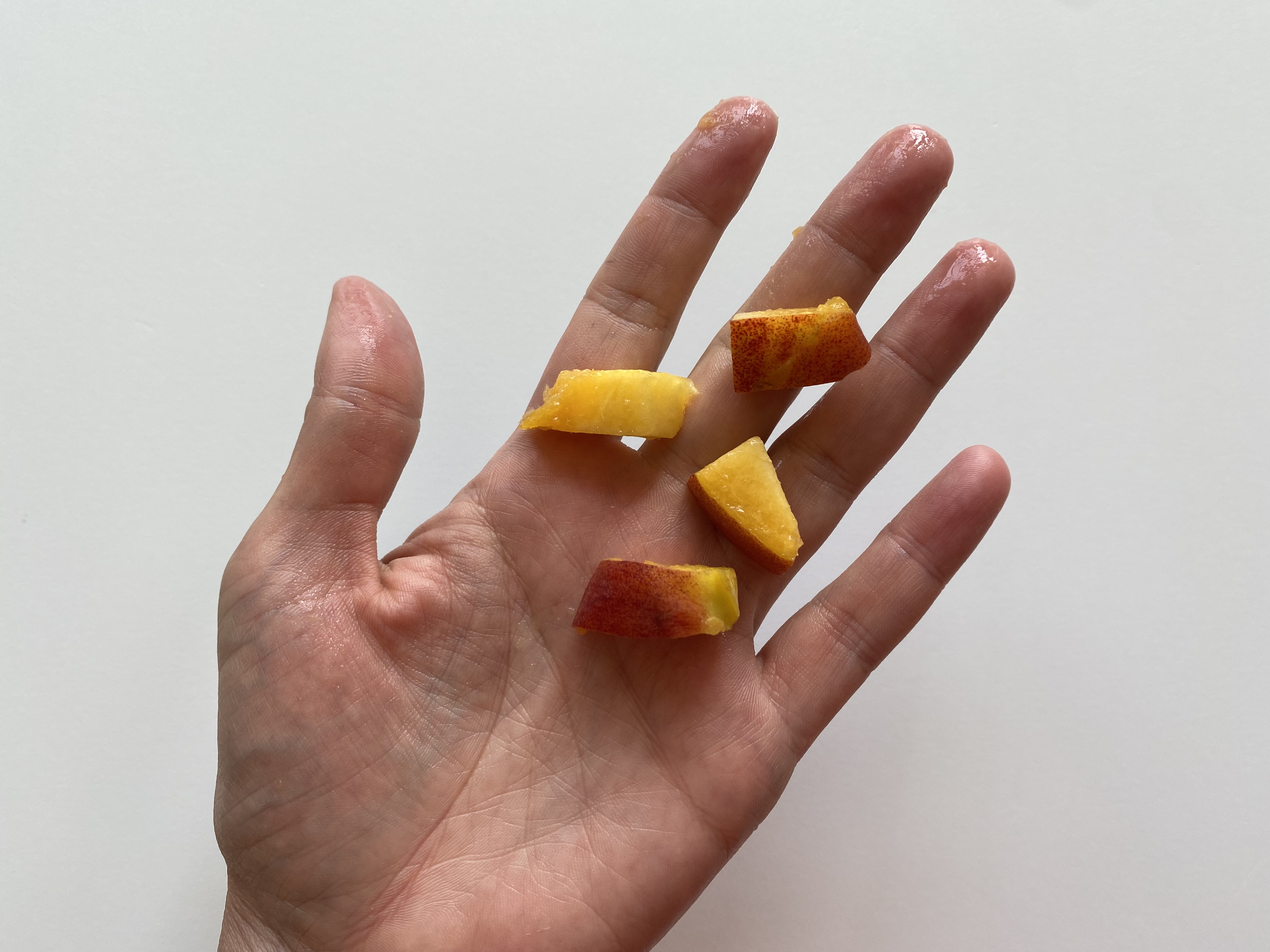 a hand holding four bite-sized pieces of peach with the skin still on for toddlers 12 months+