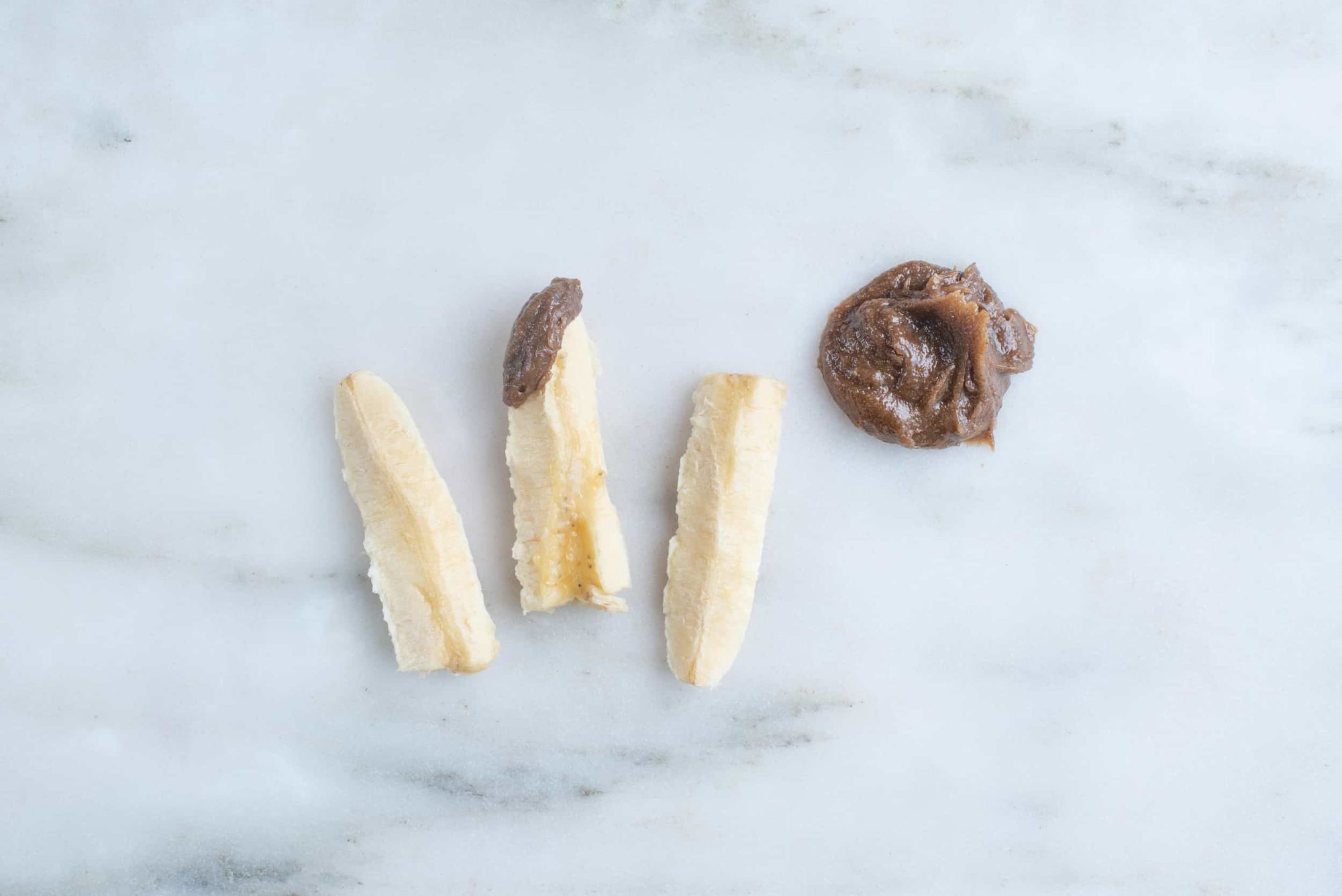 three banana spears, middle one dipped in sunbutter, sitting on a countertop next to a pile of sunbutter