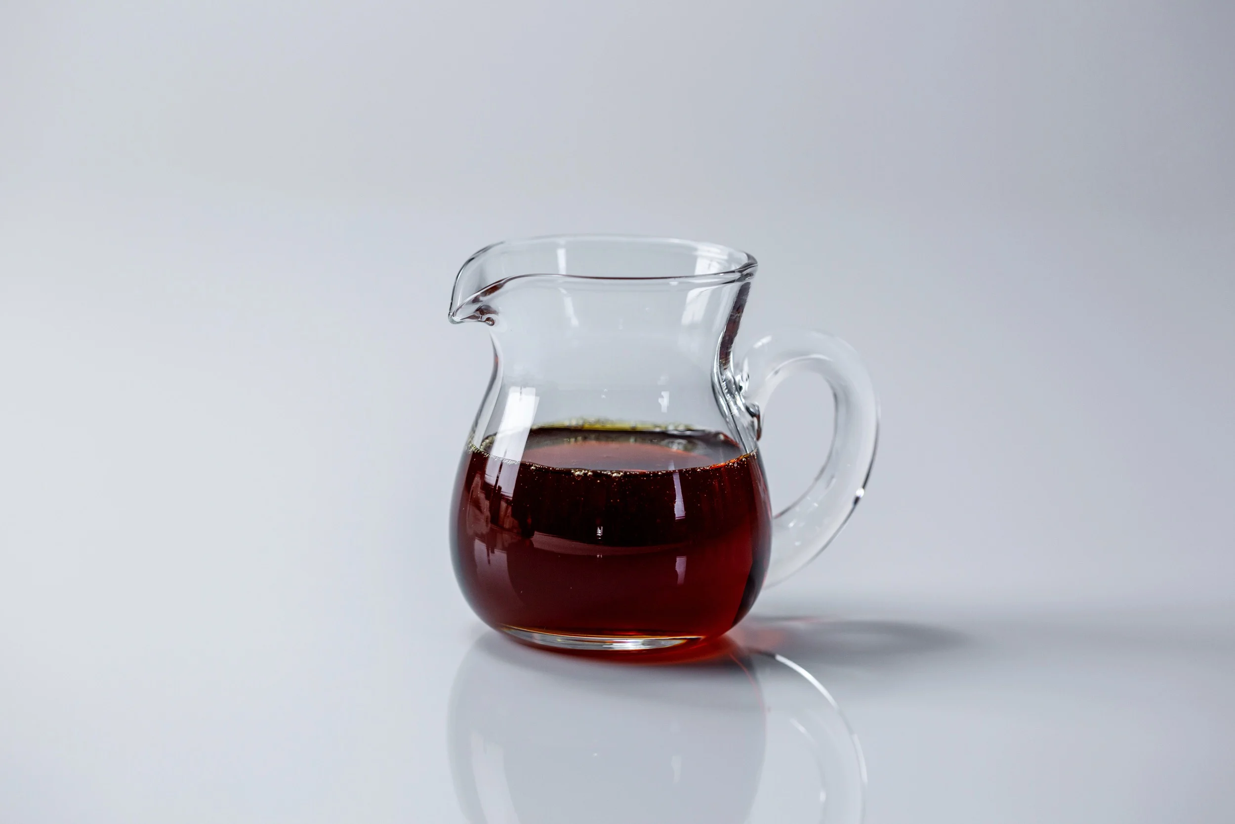 a photograph of a small clear glass pitcher filled with maple syrup on a white background