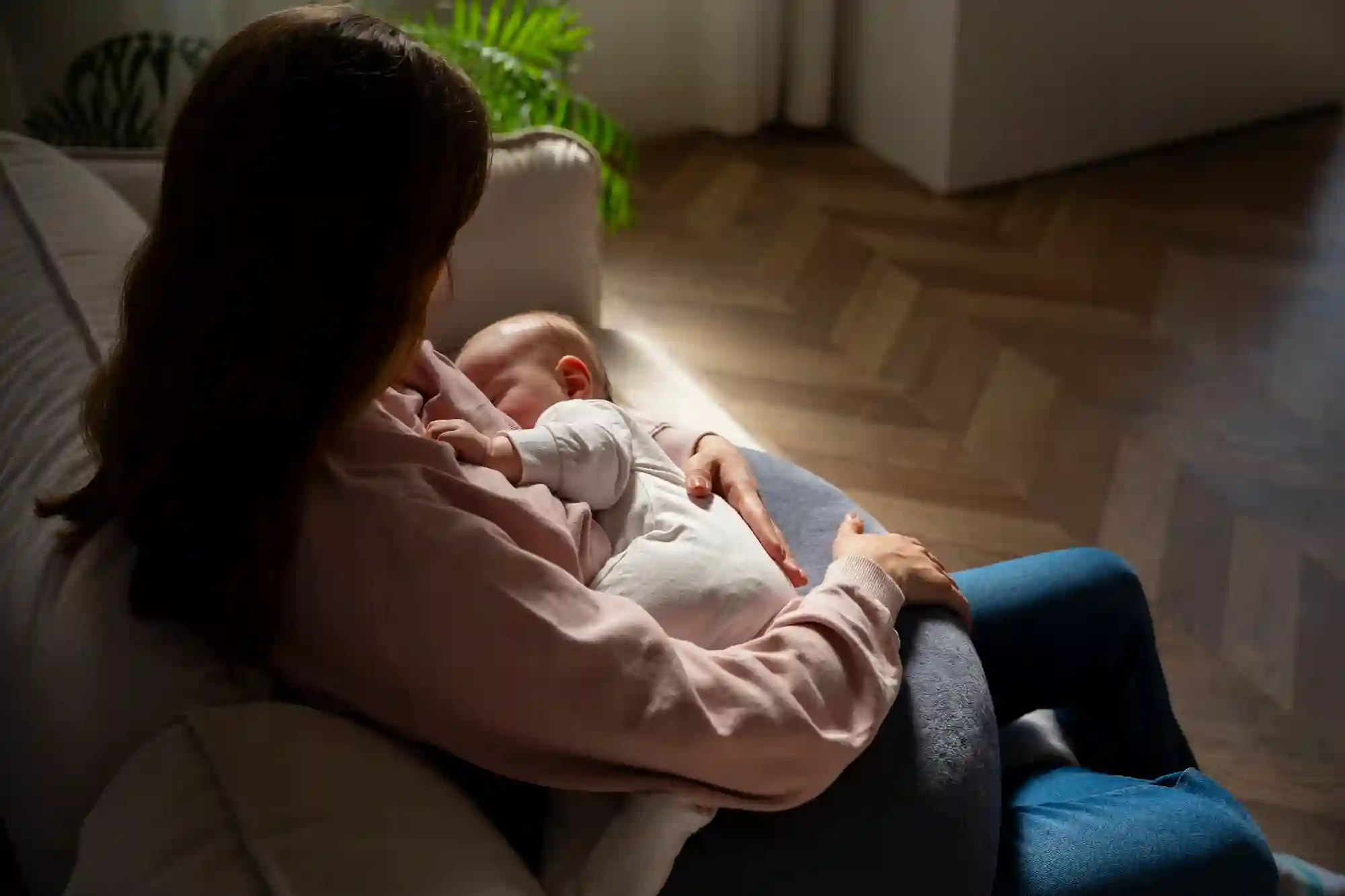 A woman in a pink sweater cradles a sleeping baby on a beige couch in a cozy, sunlit room with parquet flooring and a leafy plant.