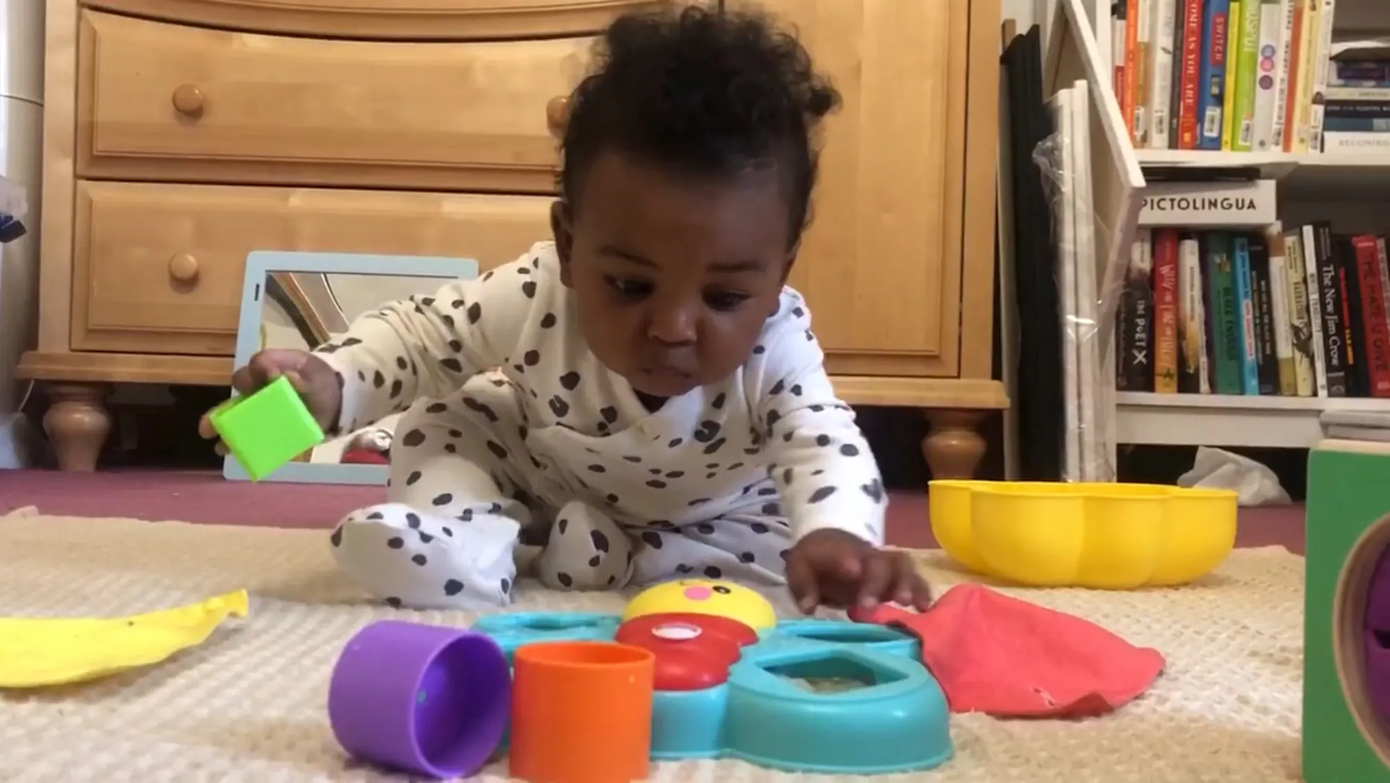 a photograph of a baby in a onesie sitting and reaching forward to grab a toy in front of him