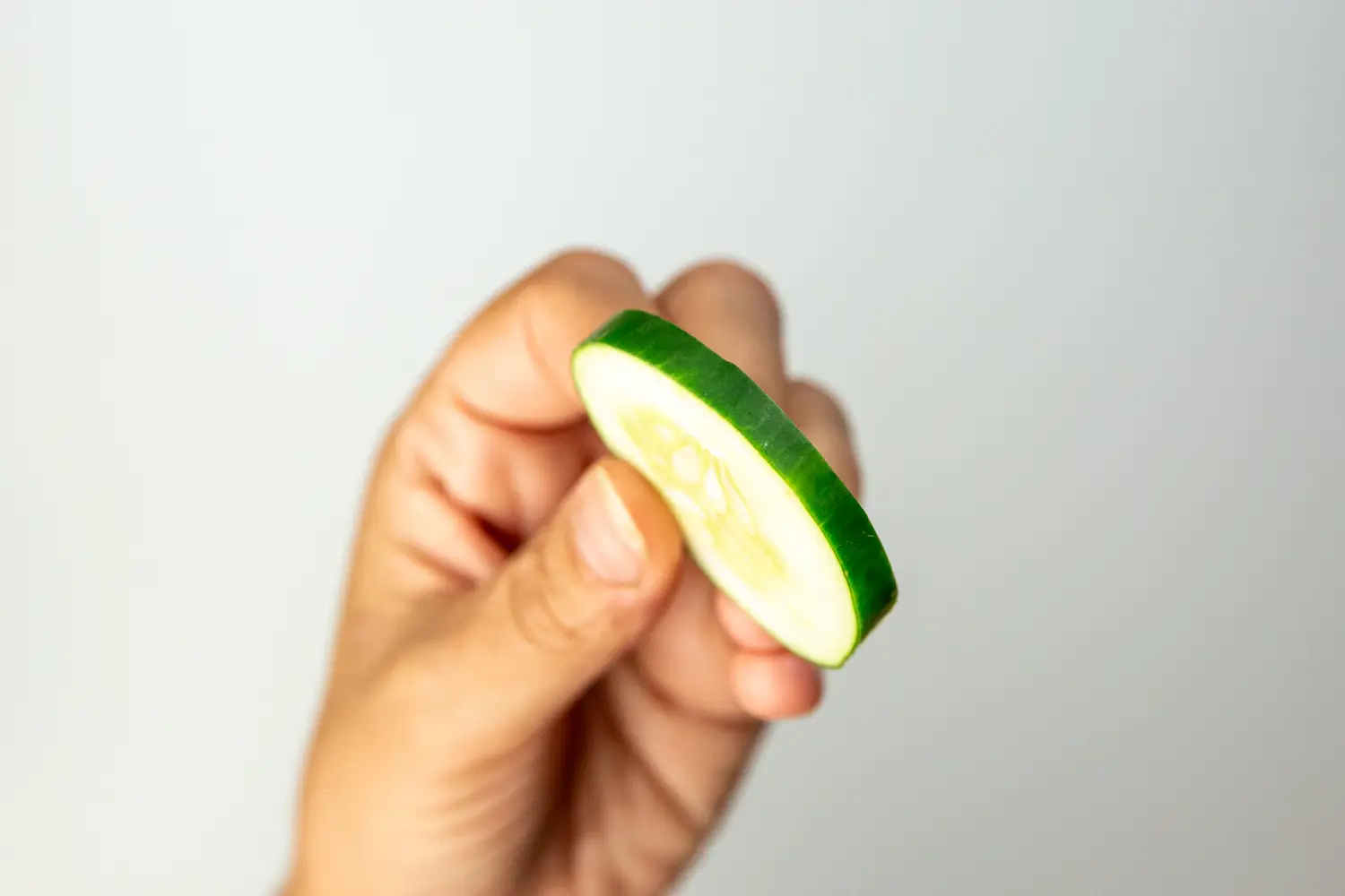 a photograph of a hand holding a thin round slice of cucumber