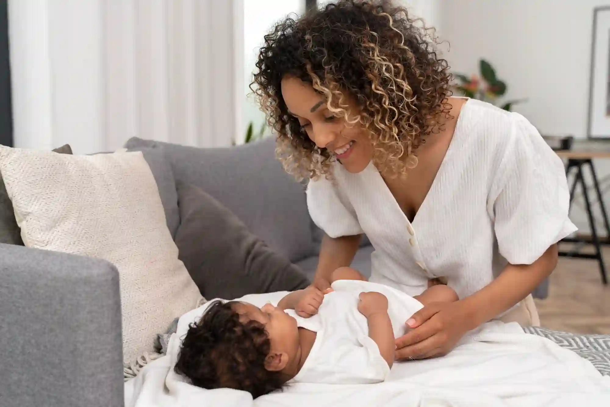 A smiling mother leans over her baby lying on a white blanket on the floor in front of a grey sofa, sharing a tender moment in a bright living room.