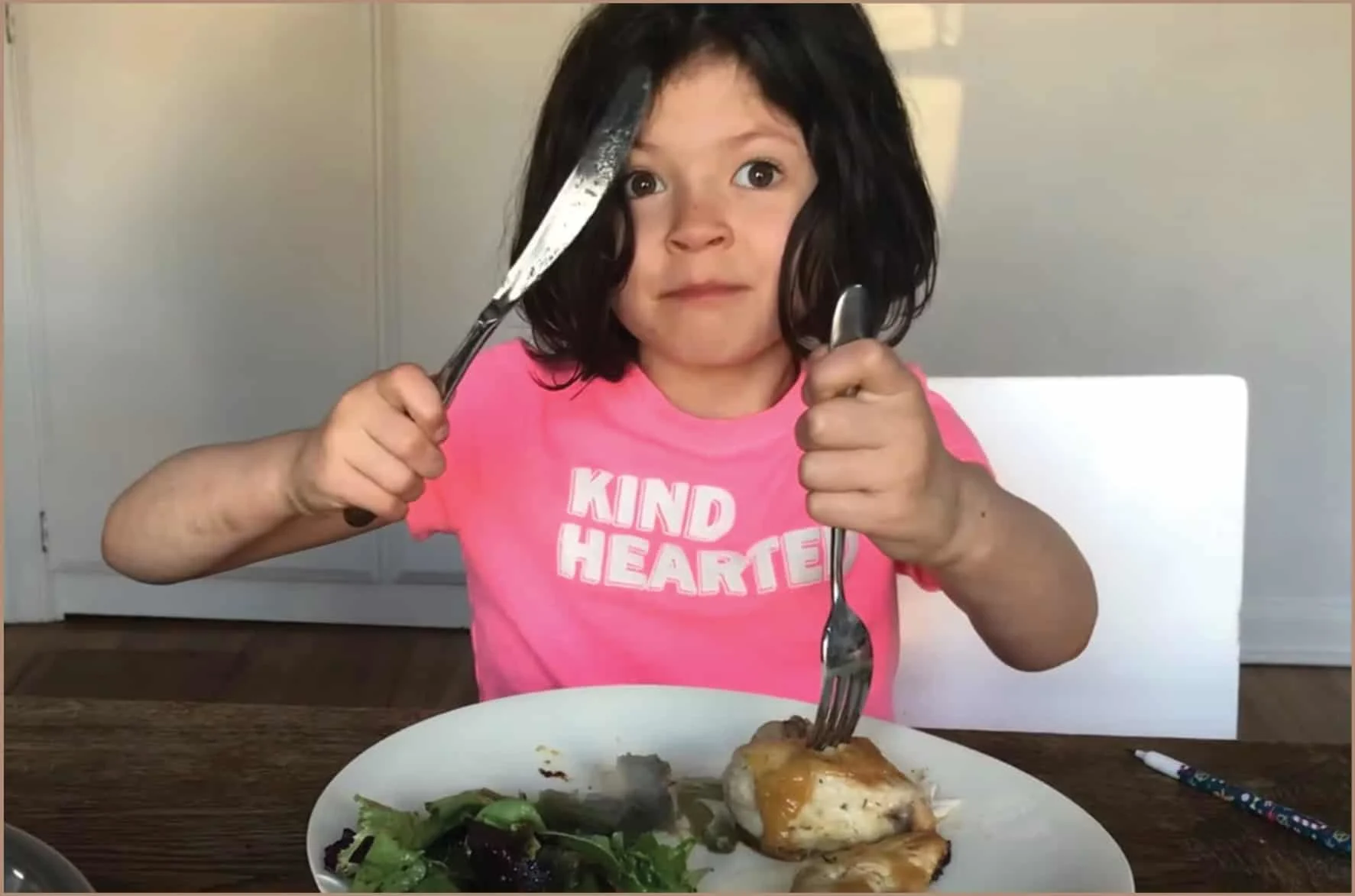 una niña pequeña sentada a la mesa comiendo, sosteniendo un cuchillo y un tenedor