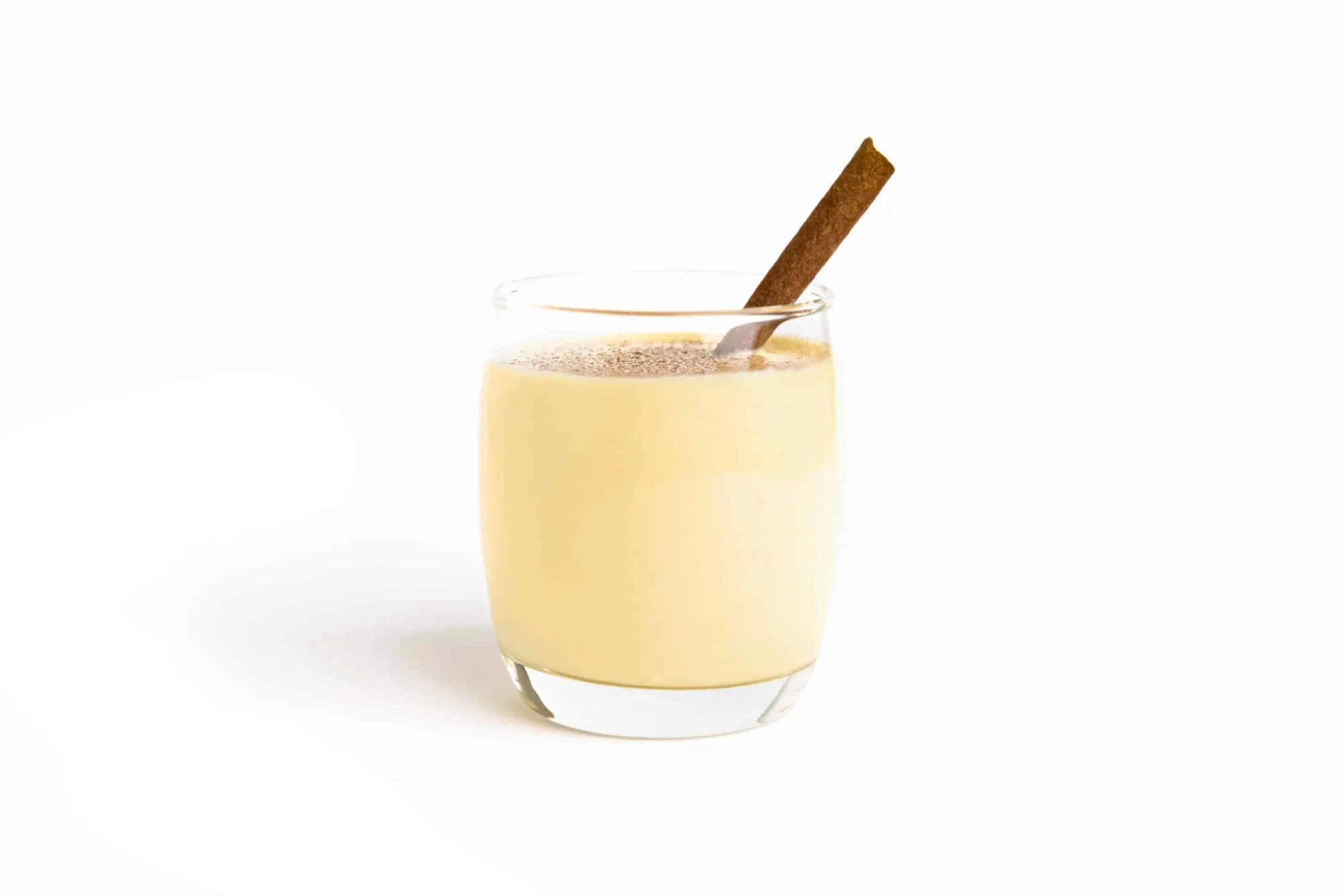 a small glass filled with creamy yellow eggnog with a cinnamon stick on a white background