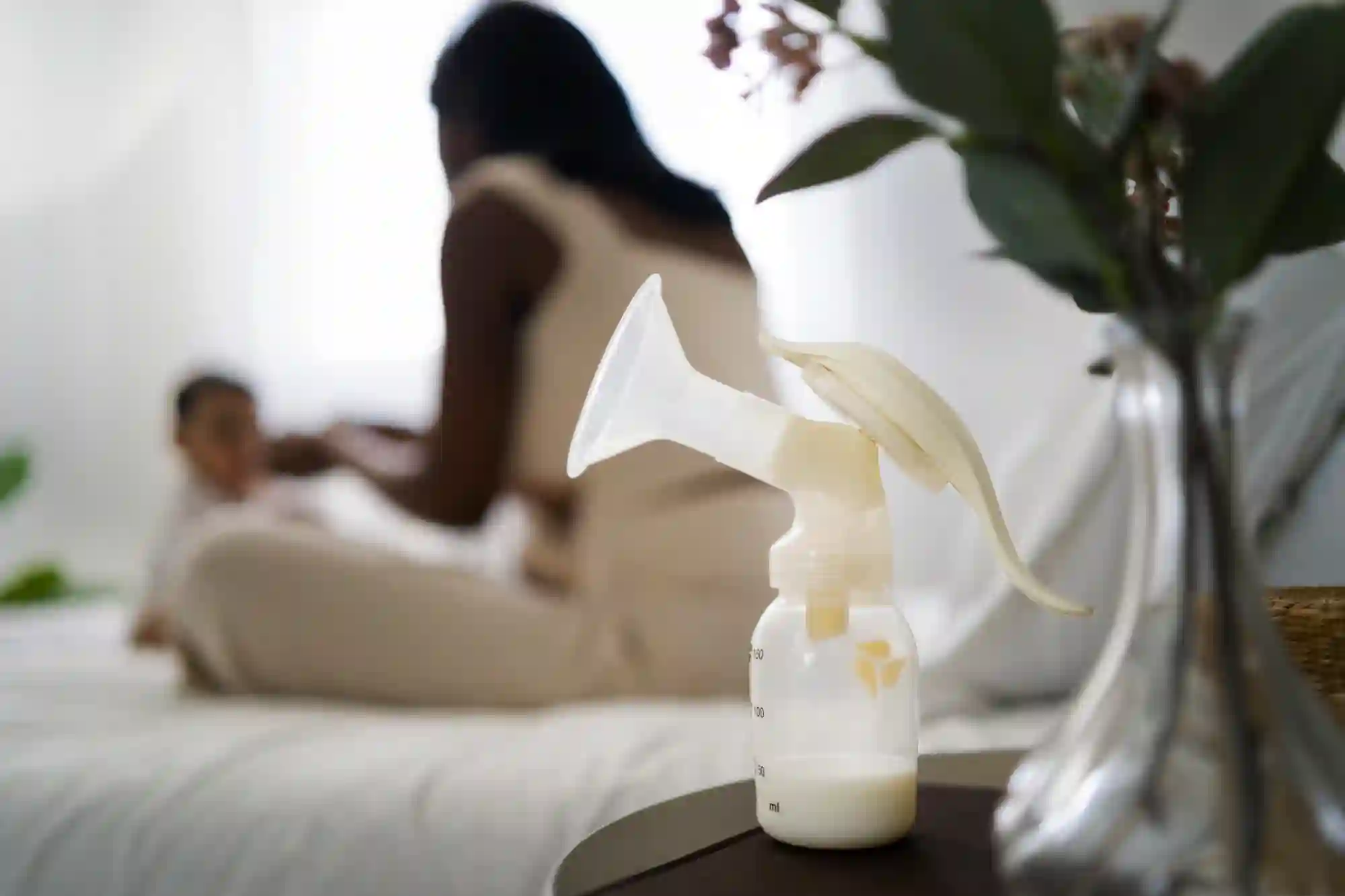 A manual breast pump with collected breast milk on a nightstand in the foreground, with a mother breastfeeding her baby on a bed in the blurred background.