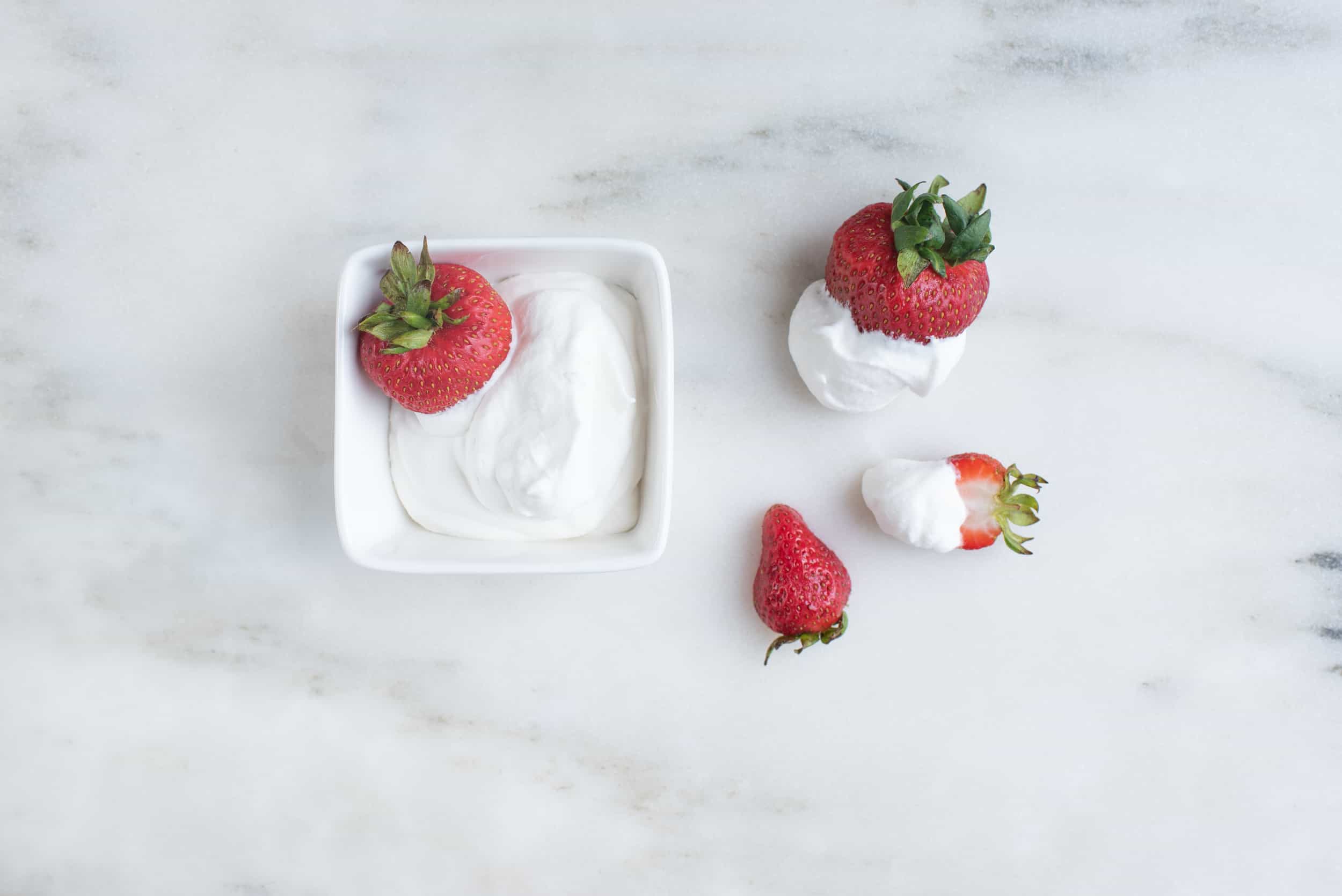 a square bowl filled with whipped cream and one whole strawberry, next to two strawberries that have been dipped in whipped cream and one small strawberry with no cream on it