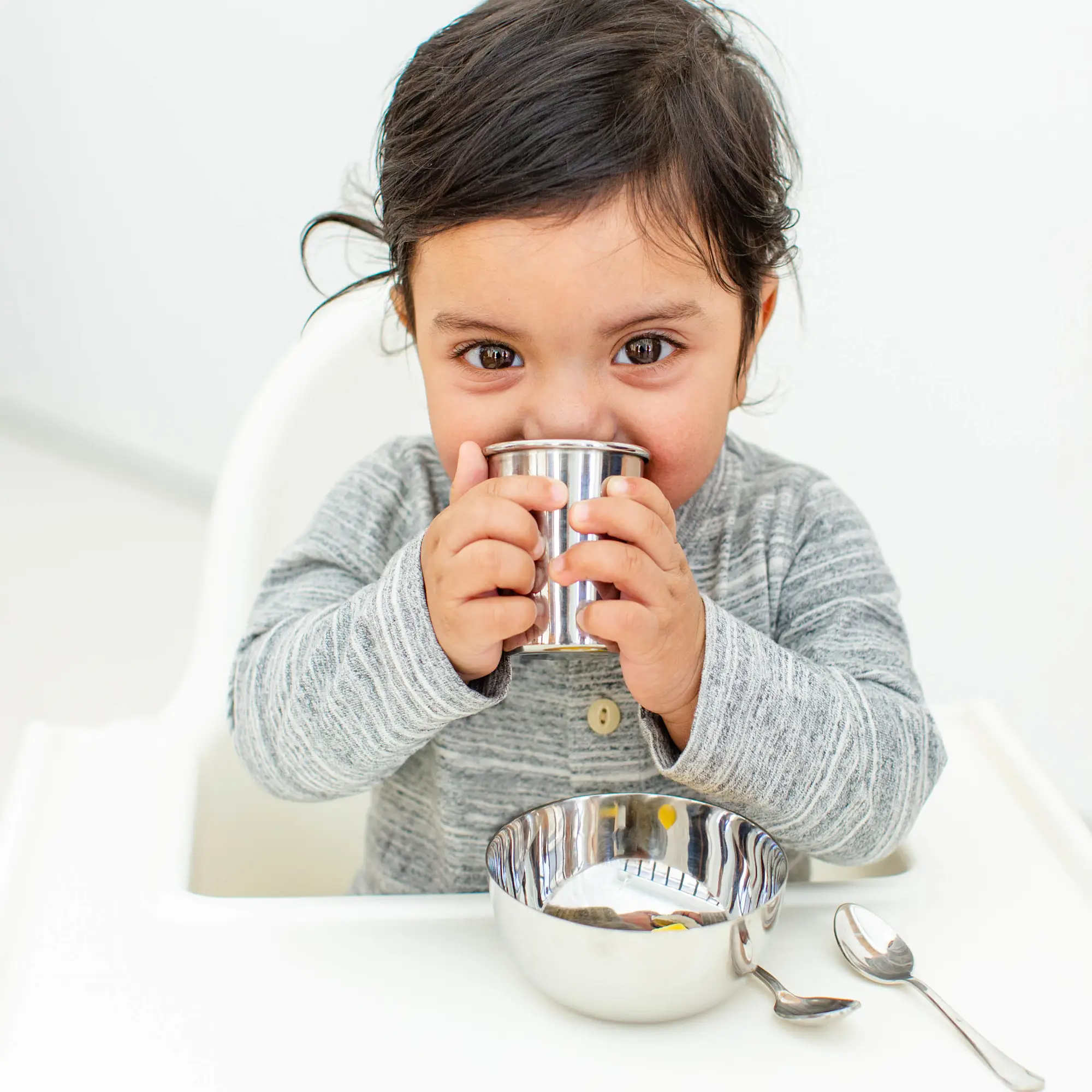 Toddler Drinking from Stainless Steel Cup A toddler in a grey sweater sitting in a white high chair, drinking from a small stainless steel cup with both hands, with a matching steel bowl and spoon on the tray in front of them.