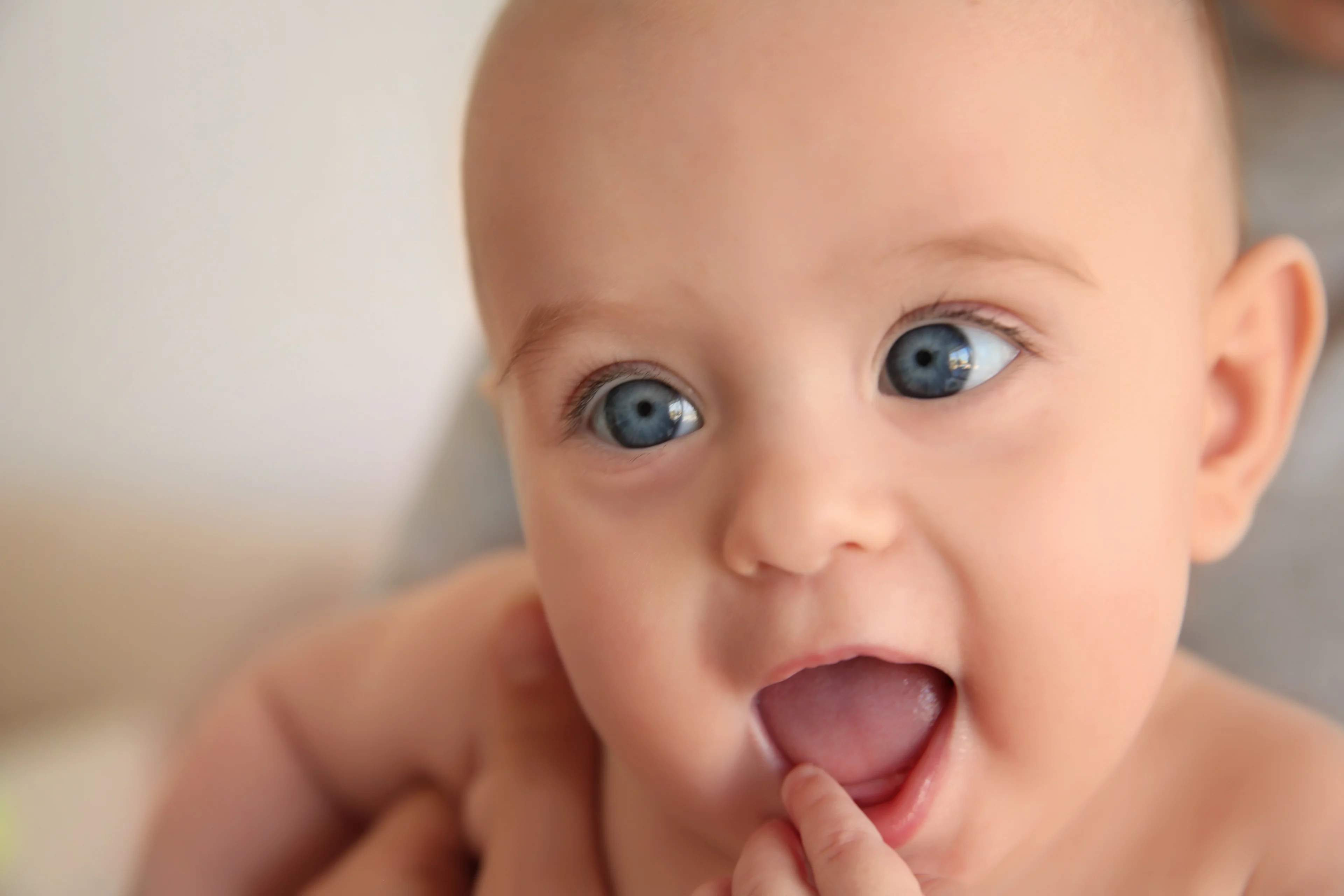 A close-up of a wide-eyed baby with bright blue eyes and mouth open, with an adult's finger touching their lower lip, against a soft blurred background.