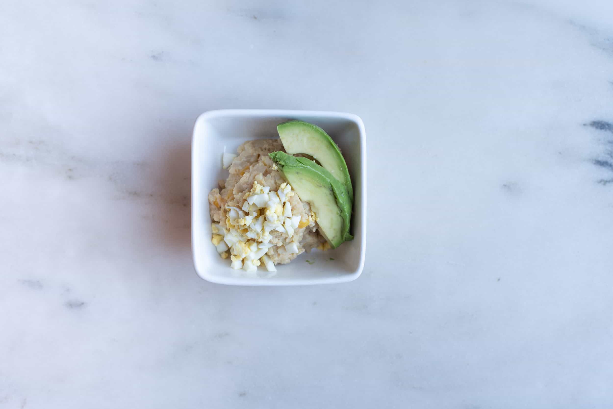 a square white bowl filled with mashed yam and two avocado slices