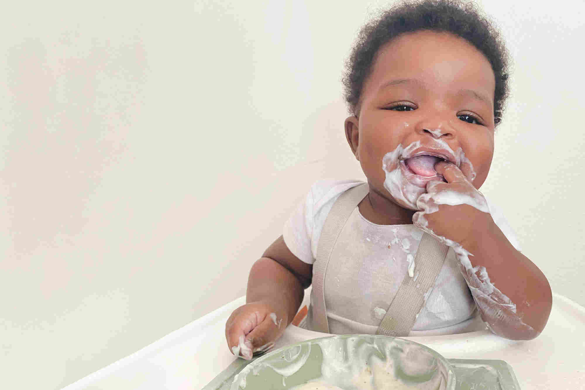 yogurt-covered baby sitting in high chair, with a finger in his mouth, with a bowl of yogurt on highchair tray 