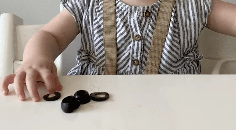 A baby in a striped top reaches toward three black olive slices on a white high chair tray, practicing the pincer grasp.