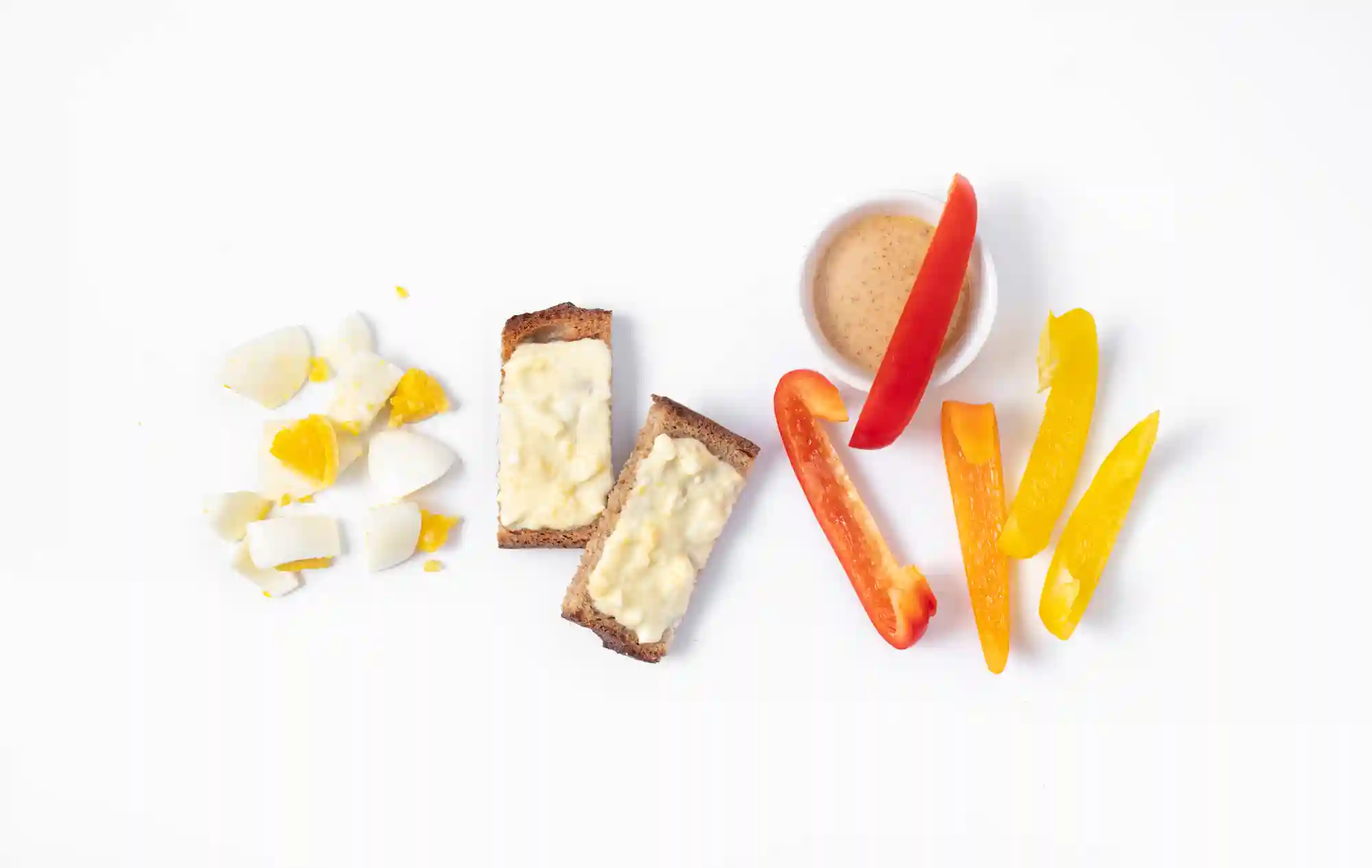A baby-friendly meal spread on a white surface featuring chopped hard-boiled egg, two slices of whole grain toast with egg spread, sliced red and yellow bell pepper strips, and a small bowl of nut butter for dipping.