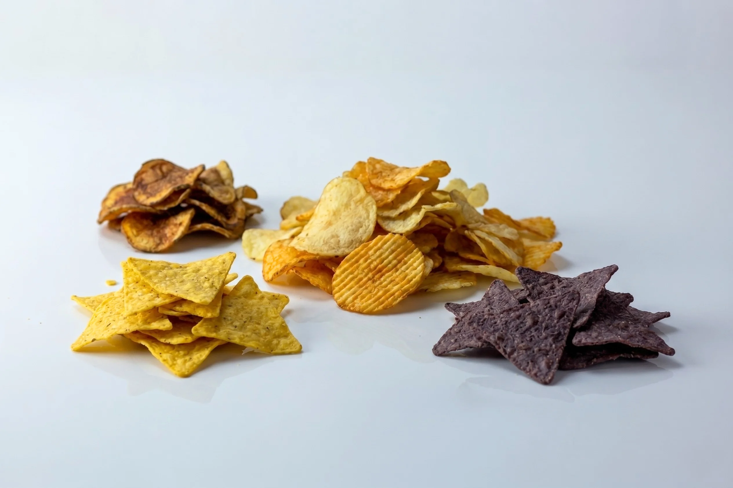 Four piles of assorted chips on a white reflective surface: a large mound of mixed potato chips in the center, yellow tortilla chips on the left, blue corn tortilla chips on the right, and darker potato chips in the back left.