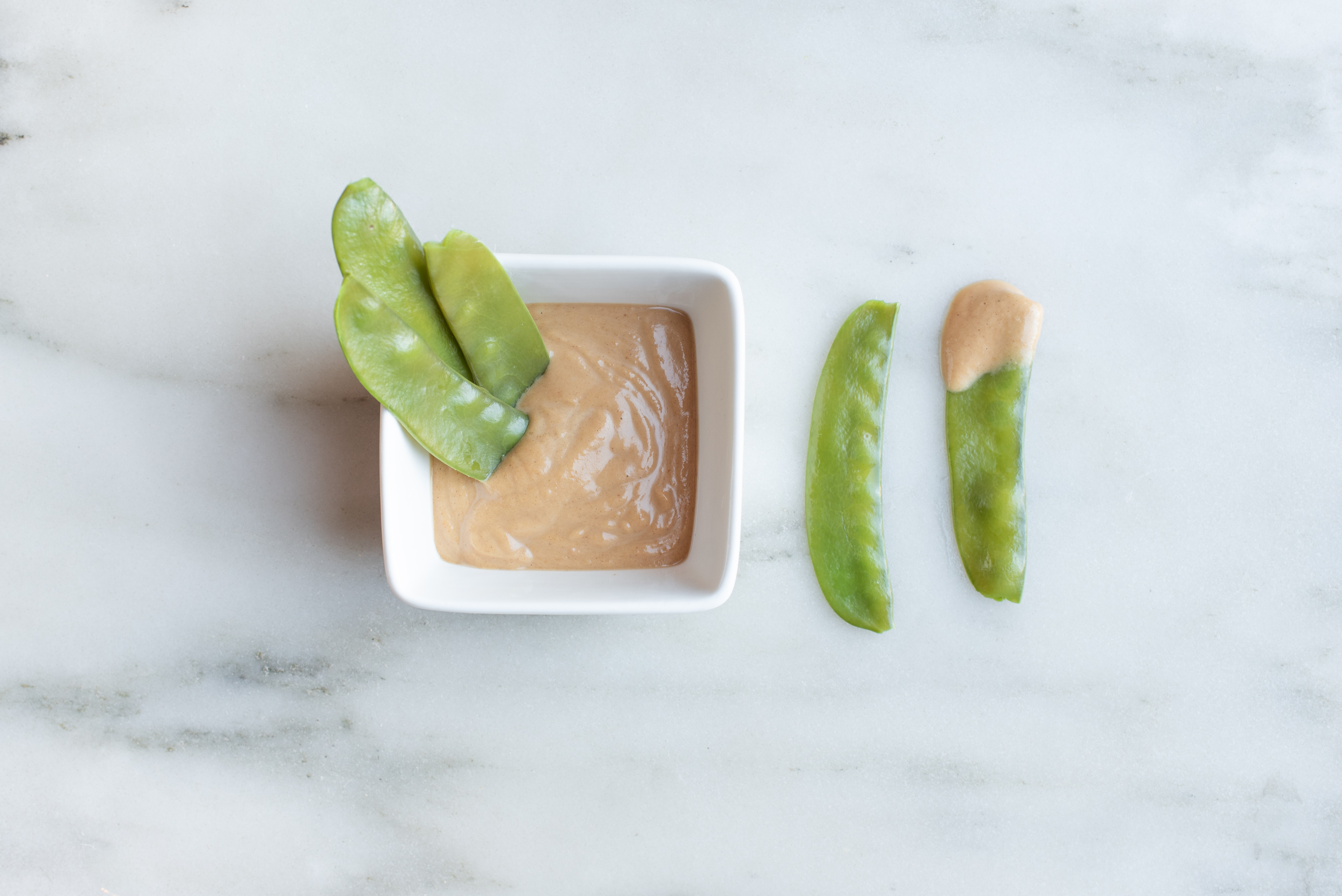a square white bowl filled with peanut butter dip with three cooked snow peas stuck in the dip next to two snow peas on a marble counter