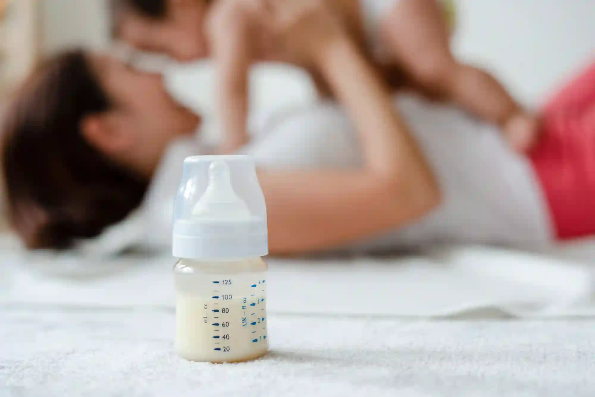 a photograph of a mom holding her baby blurred in the background with a small baby bottle with breast milk in focus in the foreground
