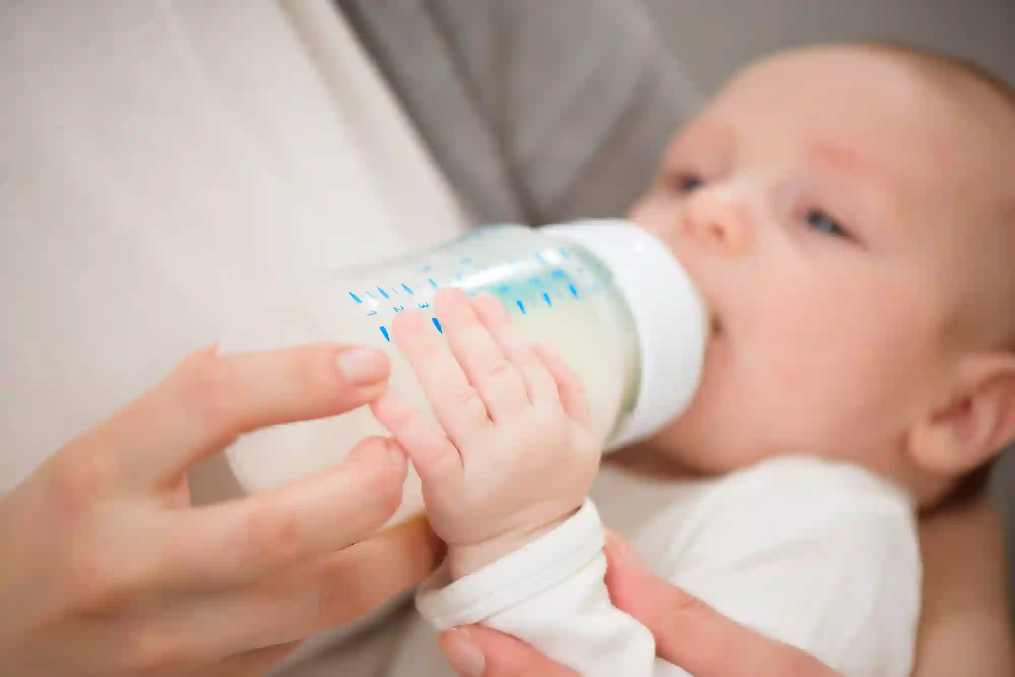 A close-up of a baby being bottle-fed milk, with the baby's small hand wrapped around the bottle alongside an adult's hand.
