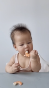 Baby holding a piece of shrimp that has been sliced lengthwise to her mouth, sitting at a table with another piece of sliced shrimp on it