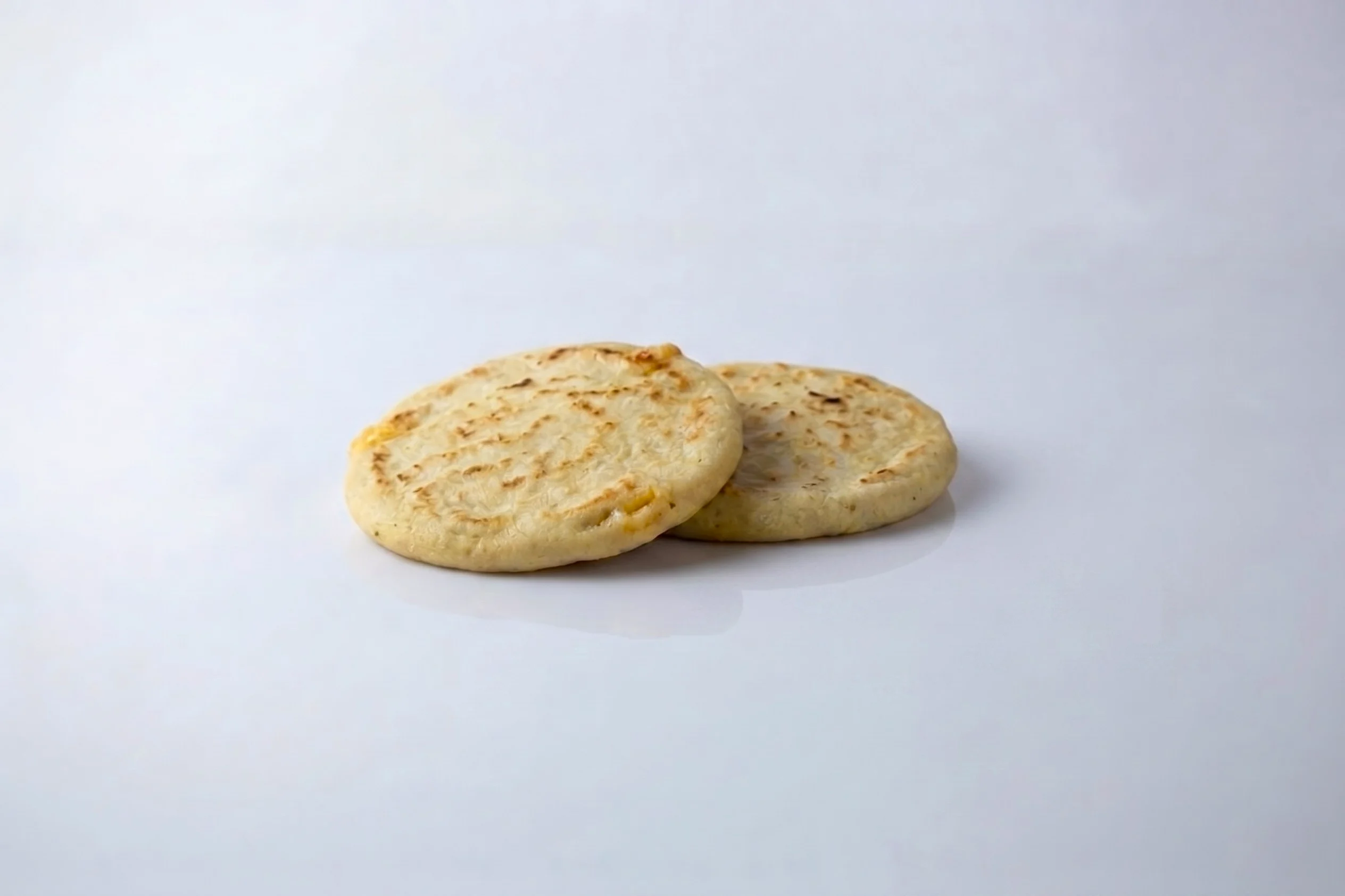 Two round, golden-griddled pupusas with lightly charred surfaces on a white reflective surface against a light grey background.