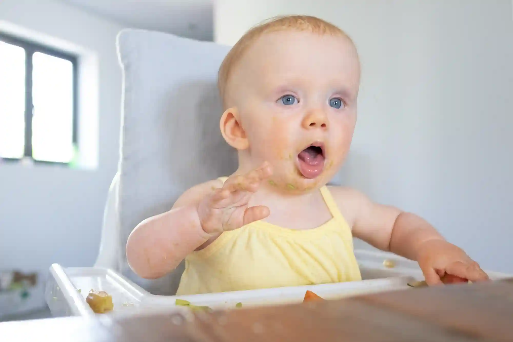 baby-gagging-on-food A blue-eyed baby in a yellow top sitting in a high chair with food smeared on her face and hands, mouth open wide, with scattered food pieces on the tray in front of her.