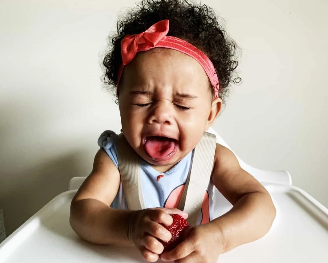 a baby in a high chair with their tongue out and eyes closed after tasting a tart strawberry