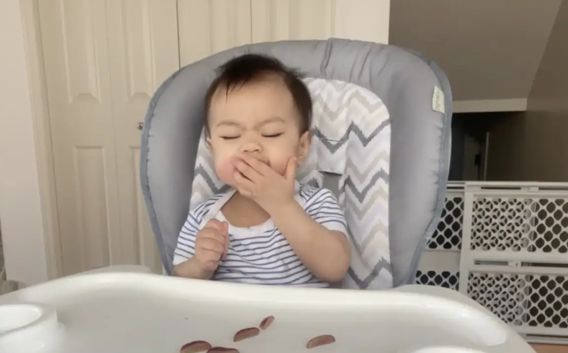 a photograph of a baby in a high chair with five pieces of quartered grape on the high chair tray and food in their mouth