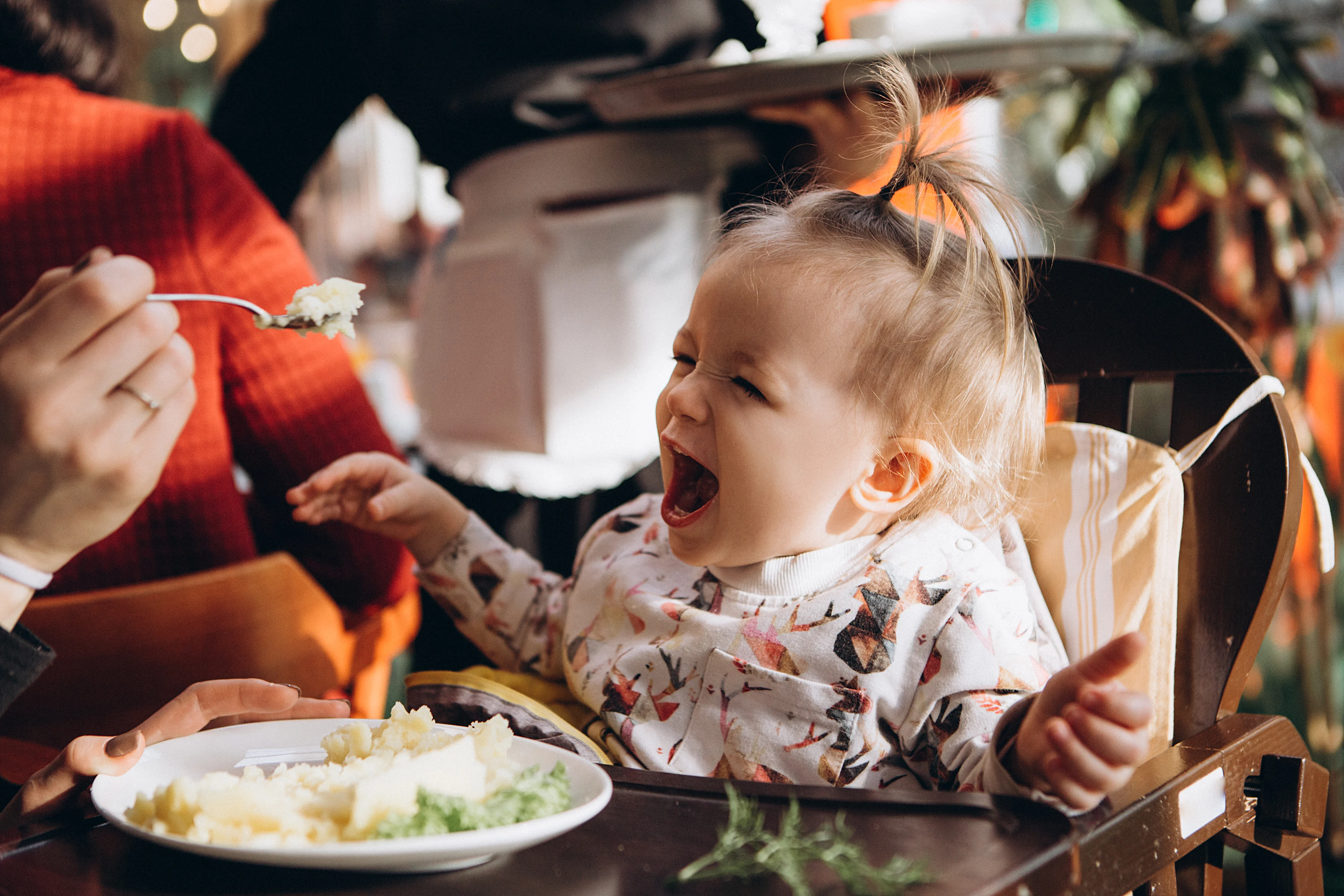 A toddler girl sitting in a high chair laughing with mouth wide open as an adult attempts to feed her mashed potatoes with a fork, with a plate of food on the tray in front of her.