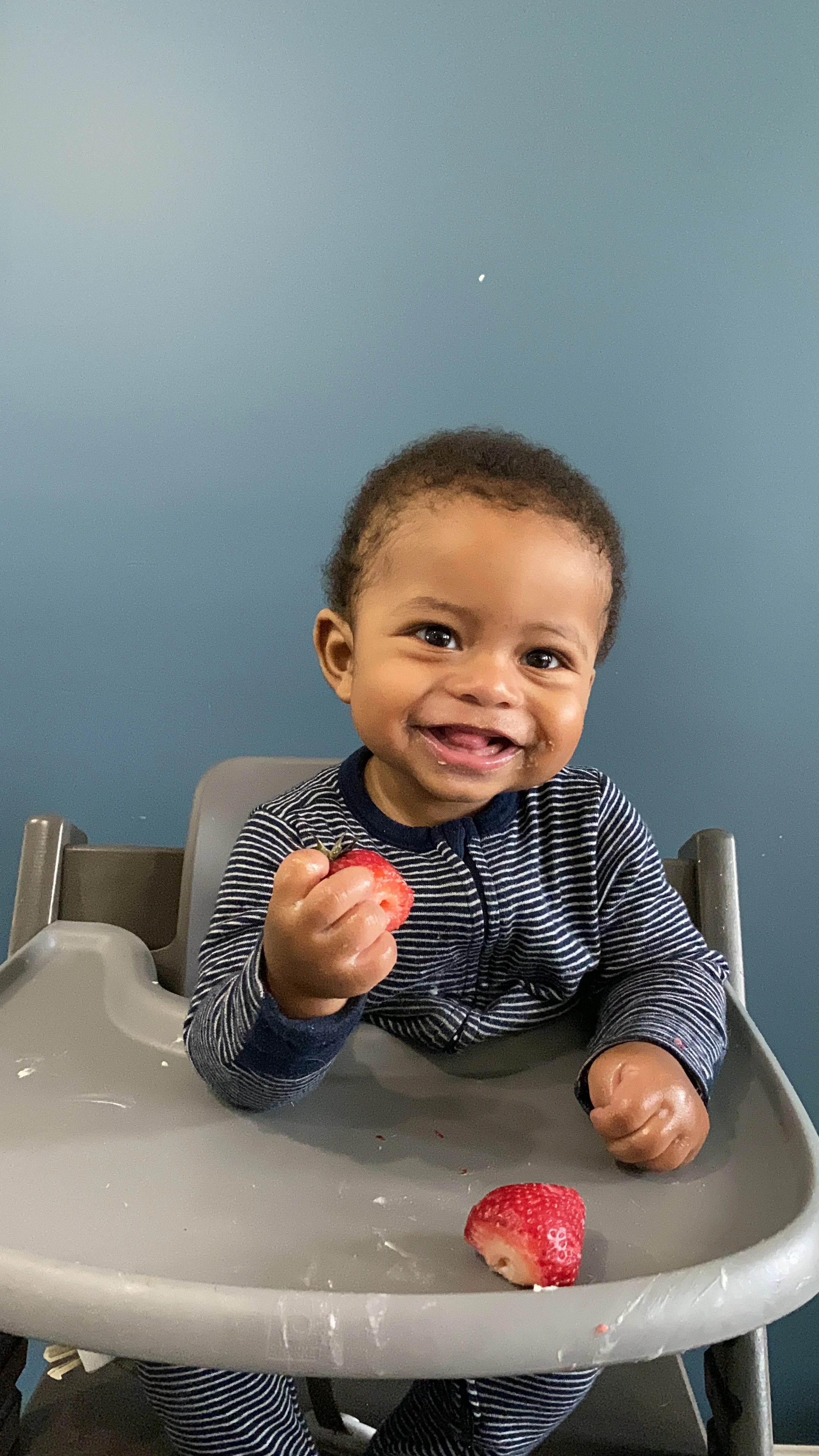 a photograph of a baby in a high chair smiling and holding a whole strawberry with another strawberry on the tray