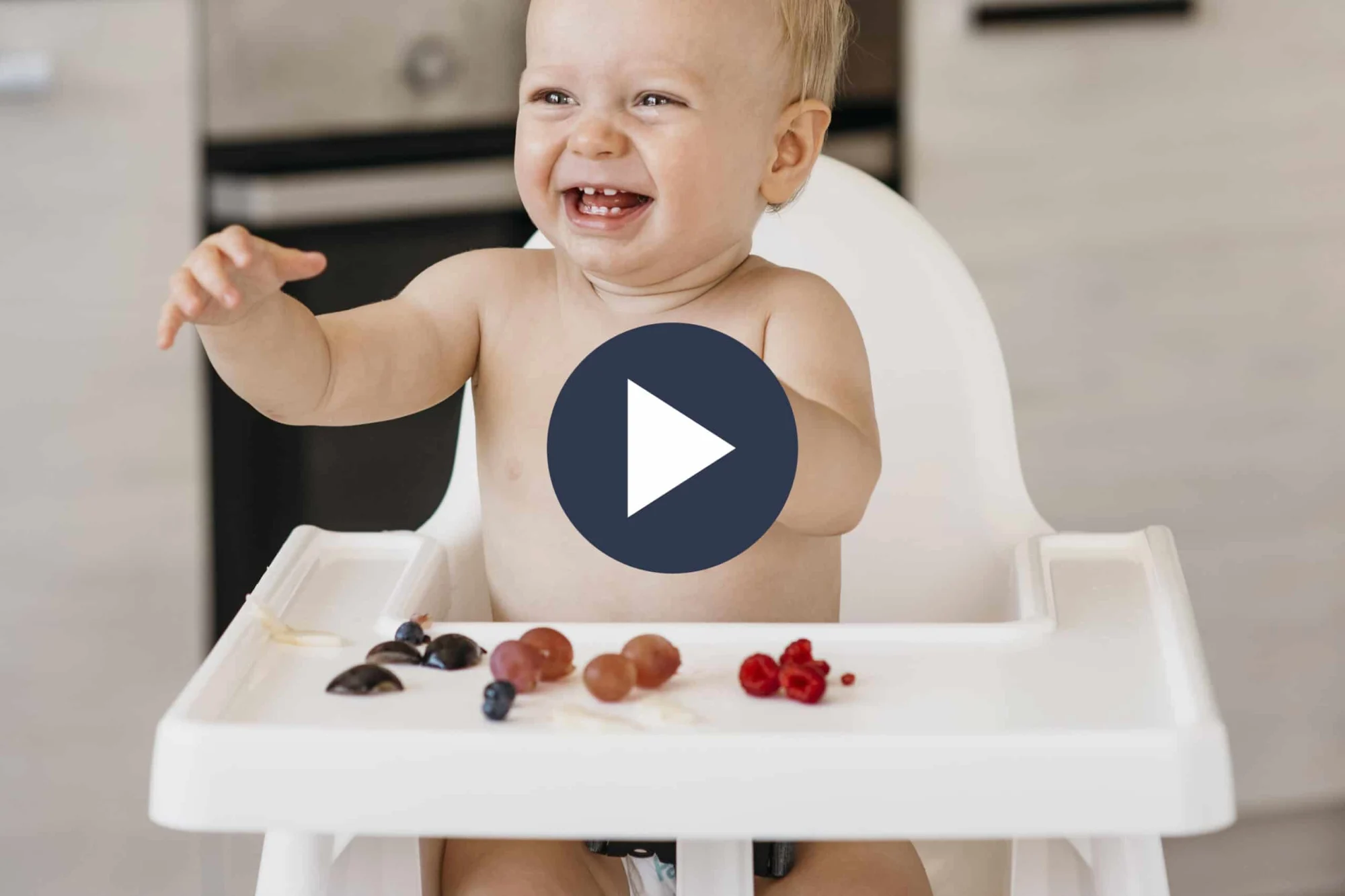 un bebé sentado en la silla alta blanca sonriendo y levantando sus brazos, con frutas cortadas en la bandeja retirable