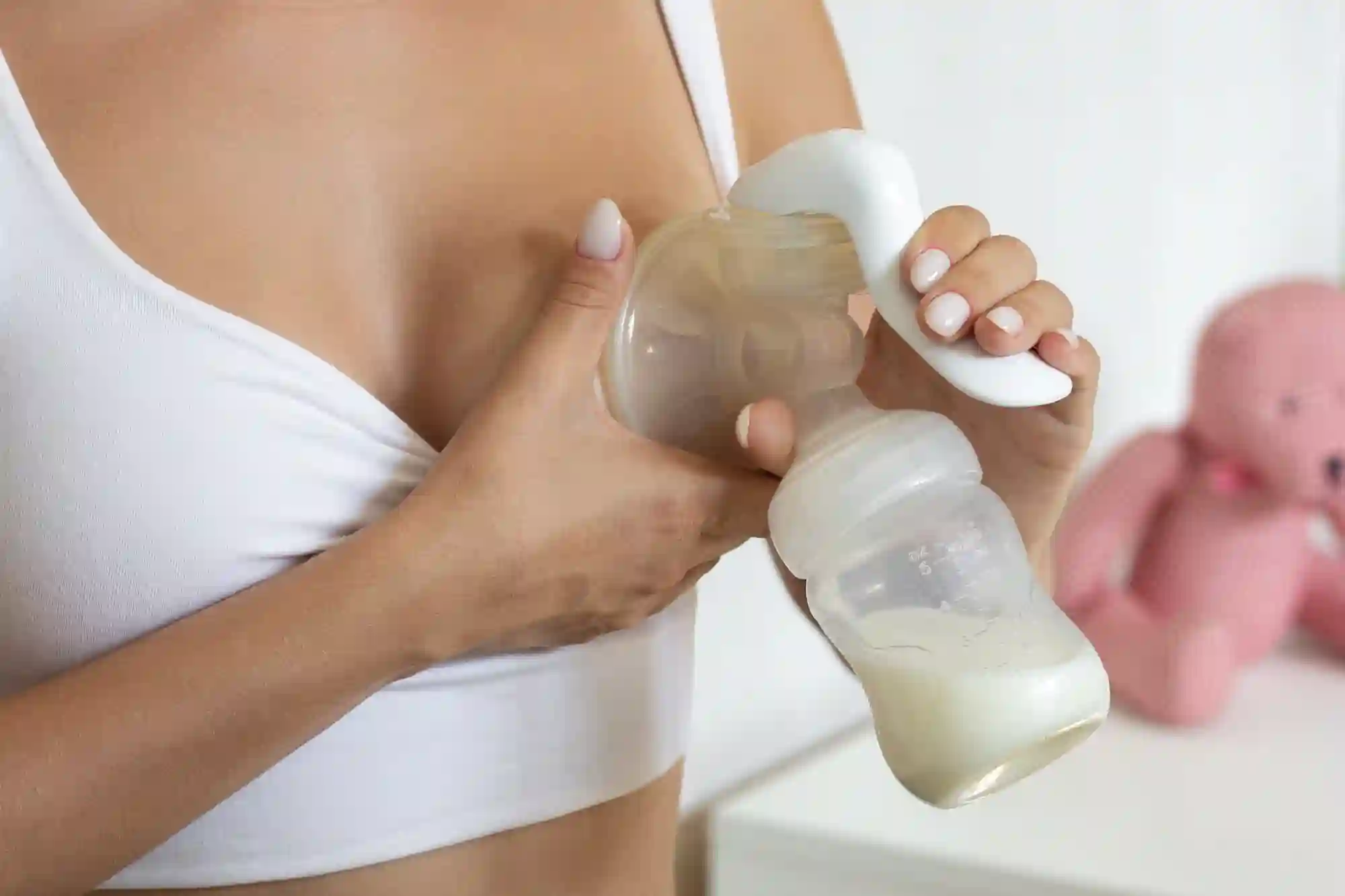 A close-up of a woman in a white nursing bra using a manual breast pump, with a bottle partially filled with breast milk, and a pink stuffed animal visible in the blurred background.
